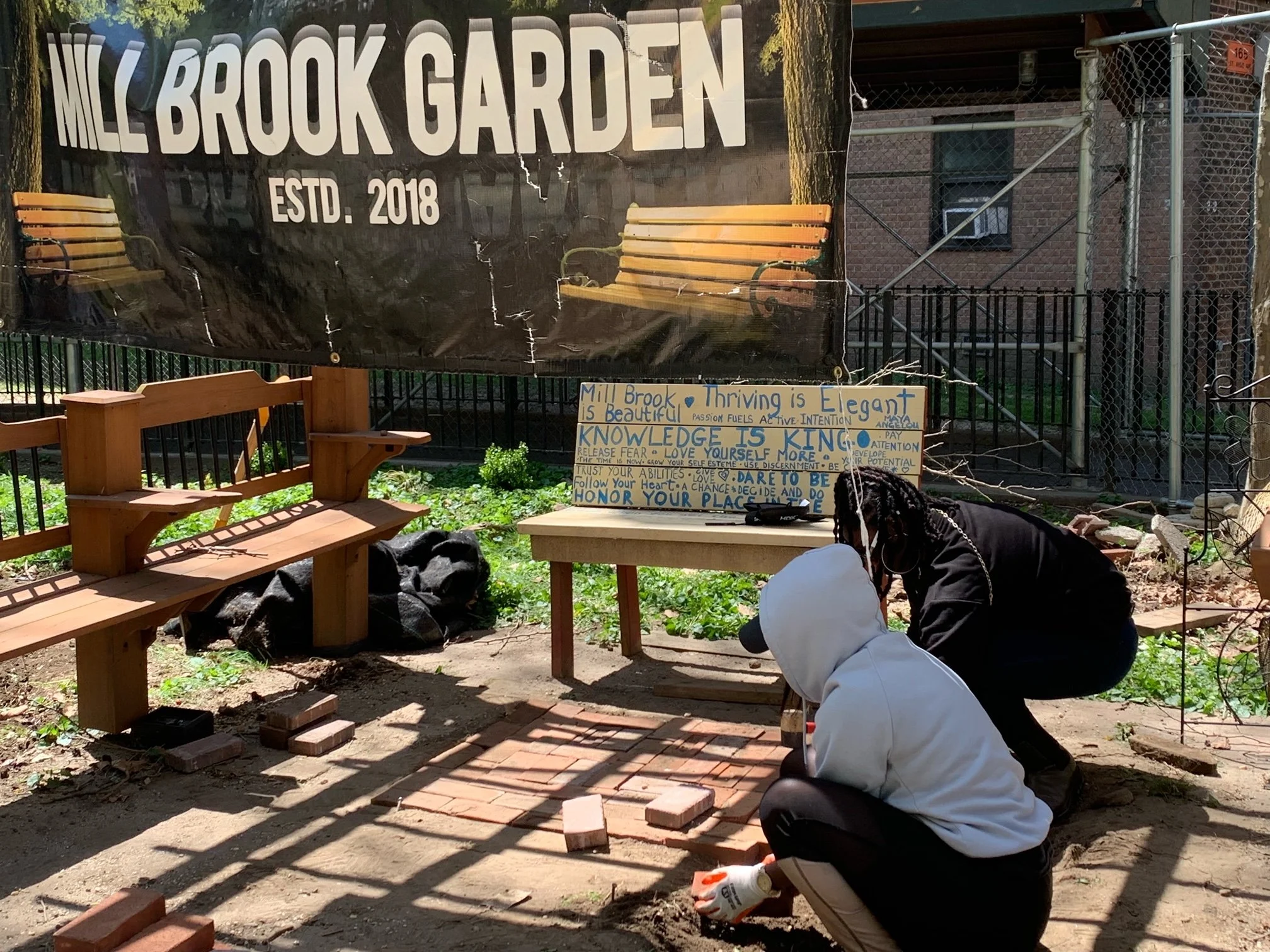 Two people are working together in a garden area with a brick patio and wooden benches. One person is squatting with glove on hand, and the other is kneeling, both focusing on laying bricks on the ground. A sign behind them reads 'Mill Brook Garden E