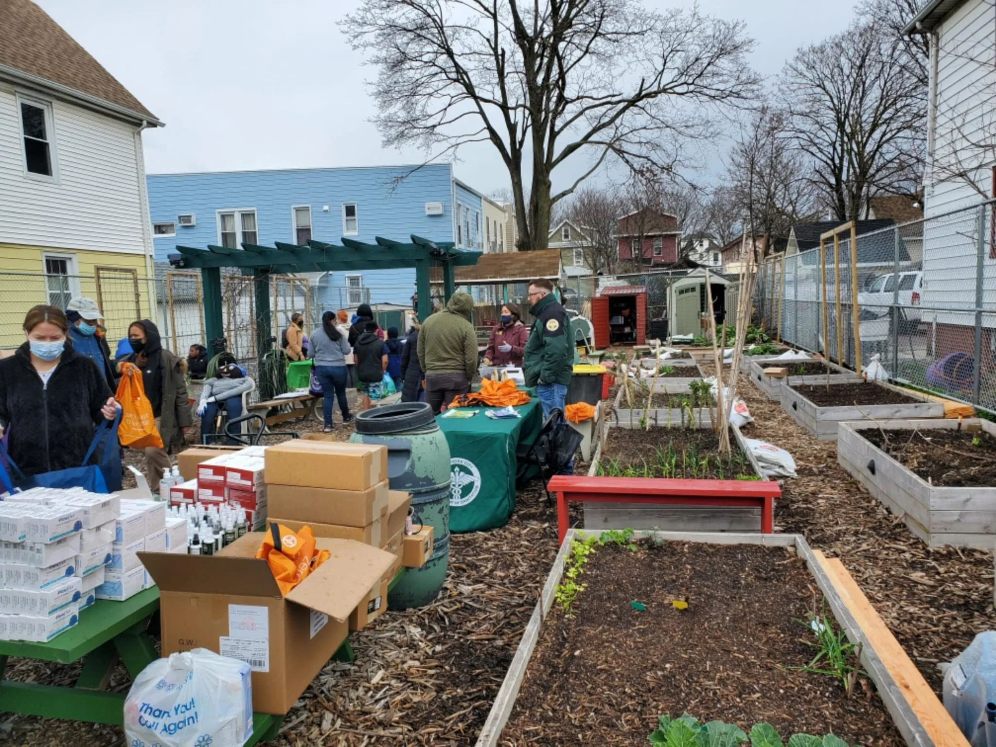 Community garden with people shopping and volunteering, vegetables planted in raised beds, tables with supplies, and a chain-link fence surrounding the area.
