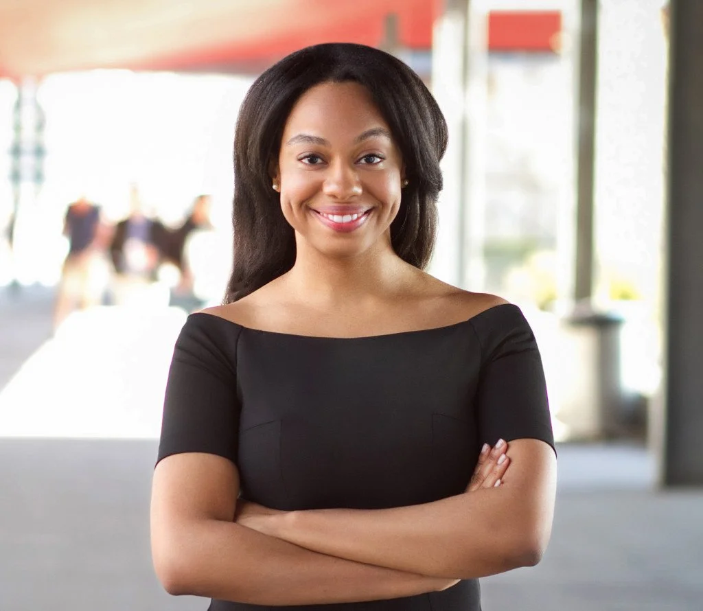 A smiling woman with medium-length black hair standing indoors with arms crossed, wearing a black top.