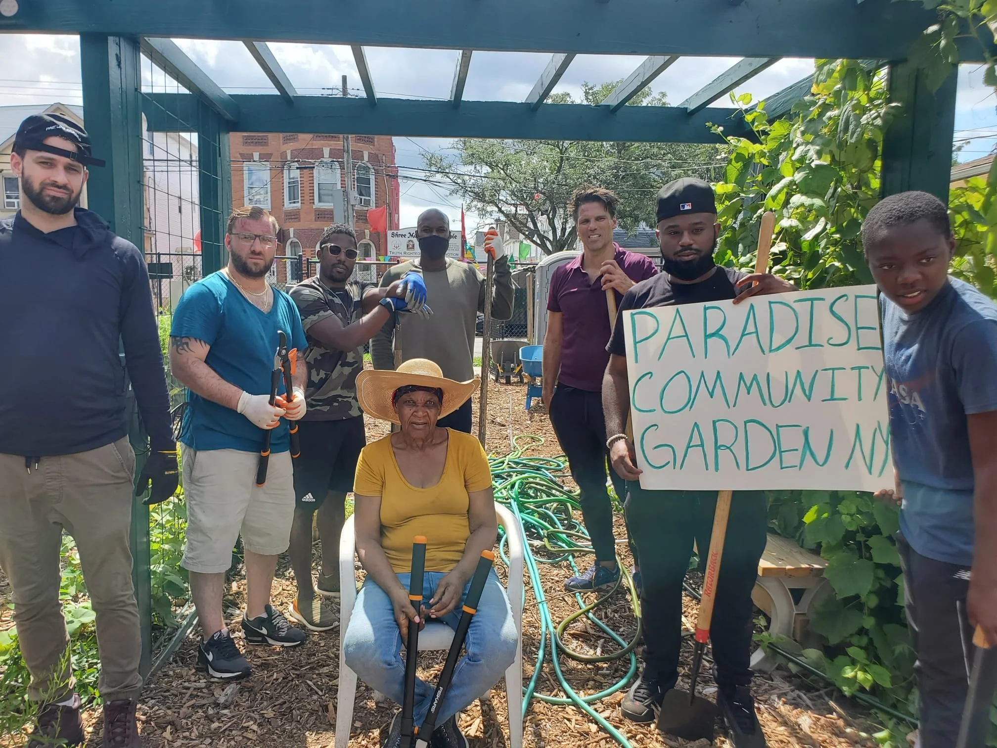 Group of diverse people standing in a community garden, holding gardening tools and a sign that reads 'PARADISE COMMUNITY GARDEN' on a sunny day.
