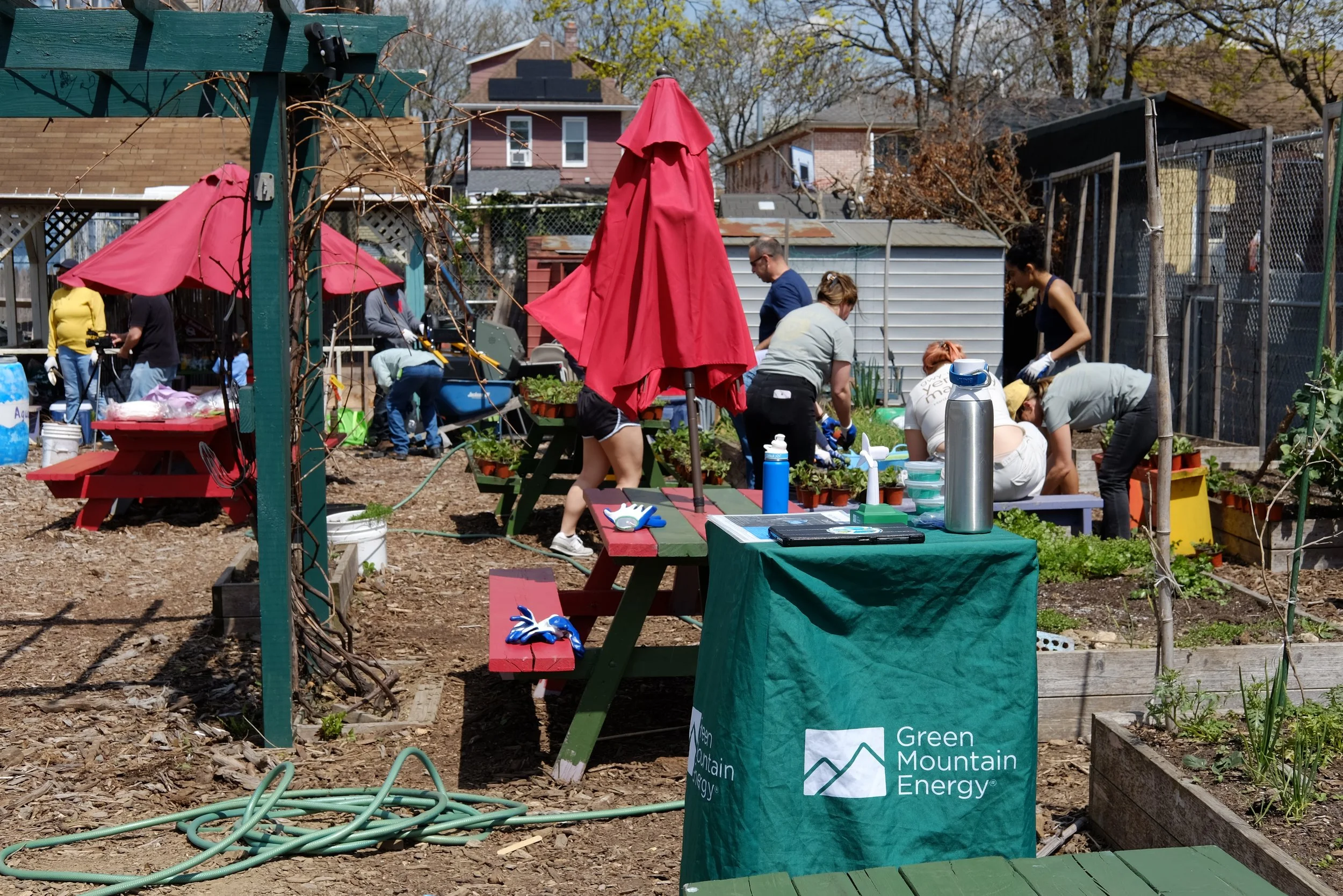 People working in a community garden, planting and tending to plants under red umbrellas, with gardening tools and supplies on tables, surrounded by a fence and houses in the background.