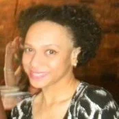 A woman smiling with short curly black hair, wearing a black and white patterned top, sitting indoors.