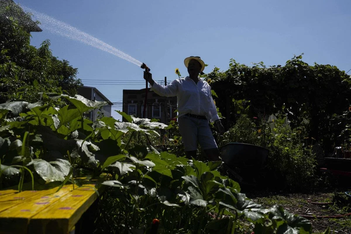 A smiling woman with long black hair, wearing a white T-shirt with colorful text, sitting outdoors on a wooden picnic table next to a pink wheelbarrow filled with yellow and purple flowers. She is holding gardening gloves.
