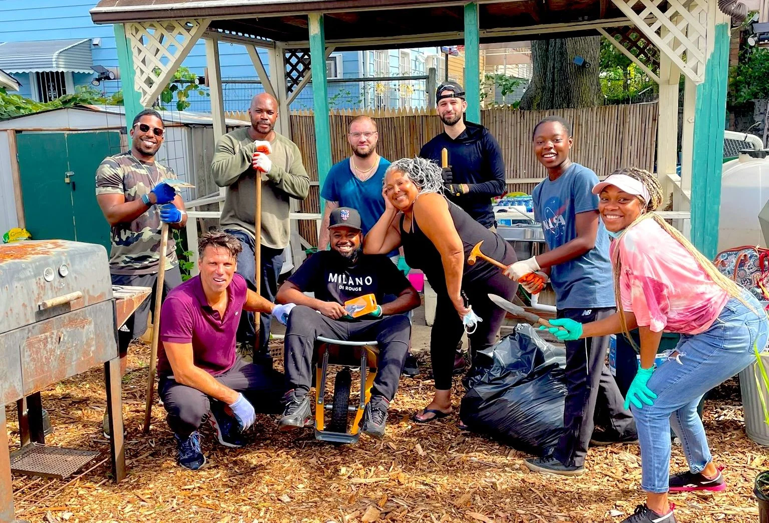 Group of diverse volunteers working together in a backyard community project, some holding gardening tools, others wearing gloves, with a wooden structure and trees in the background.