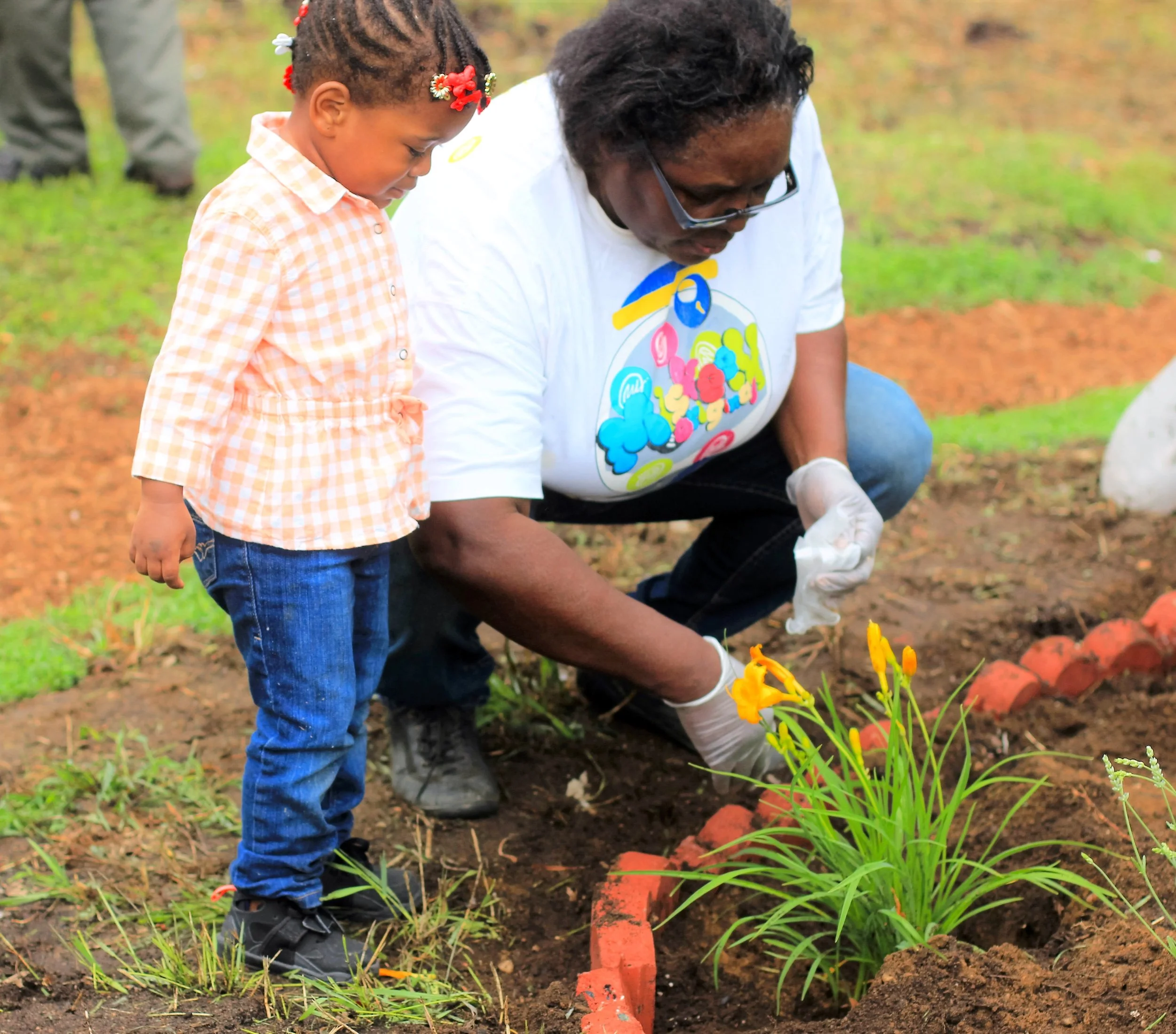 An adult and young girl planting yellow flowers in a garden bed surrounded by small red bricks. The girl has braided hair with red flower accessories. The adult is wearing glasses and a colorful graphic t-shirt.
