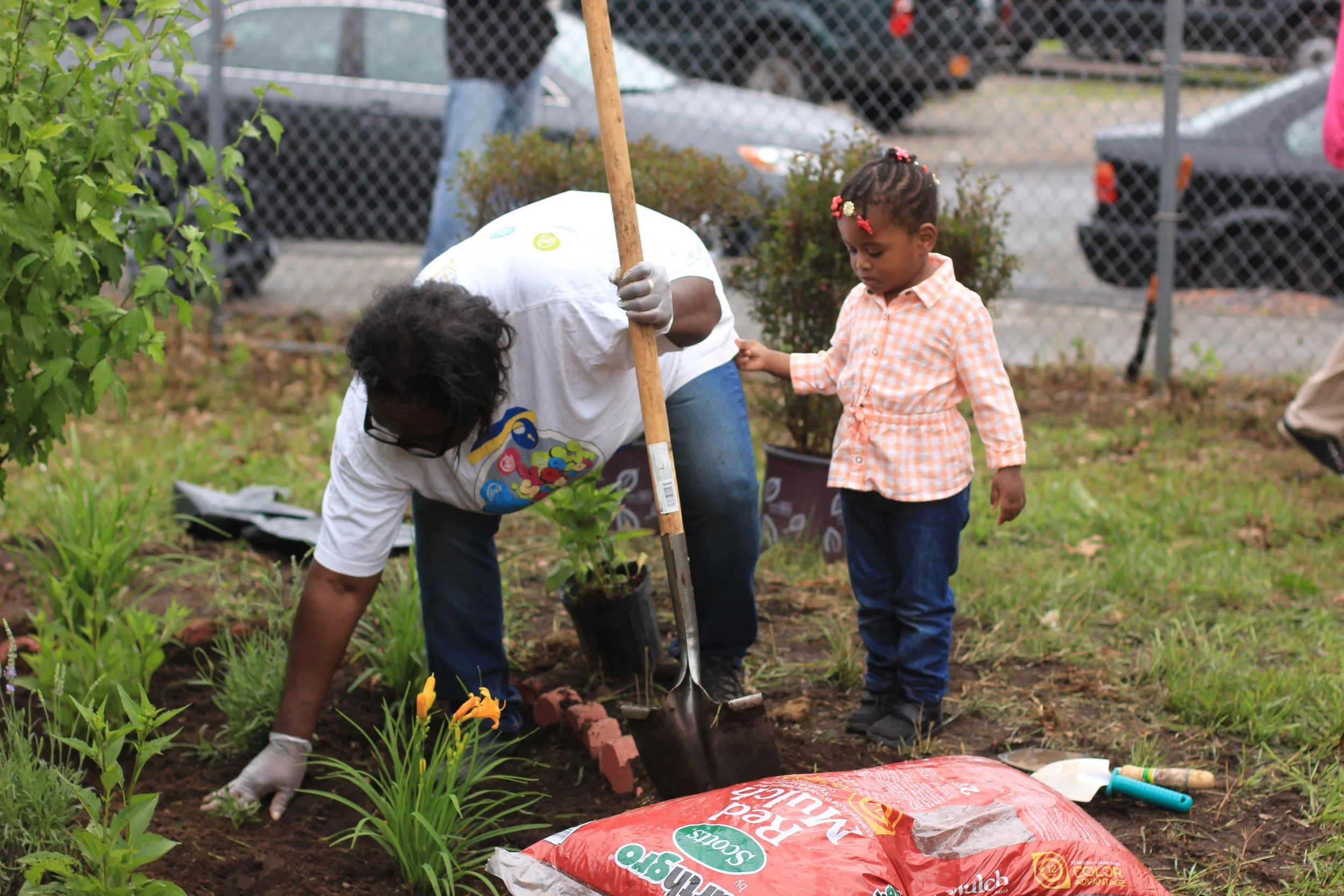 A person planting flowers in a garden with a young girl observing.