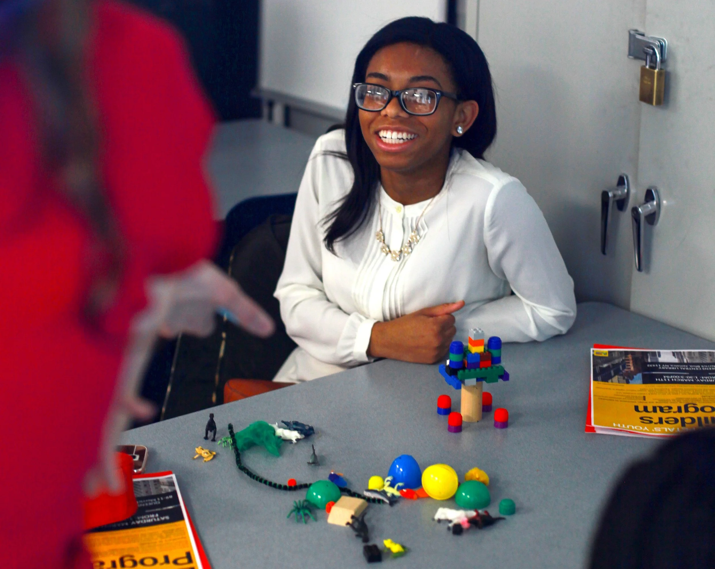 A woman with glasses, wearing a white blouse and a necklace, is sitting at a table with toys and pamphlets. She is smiling and looking at someone in red clothing in front of her.