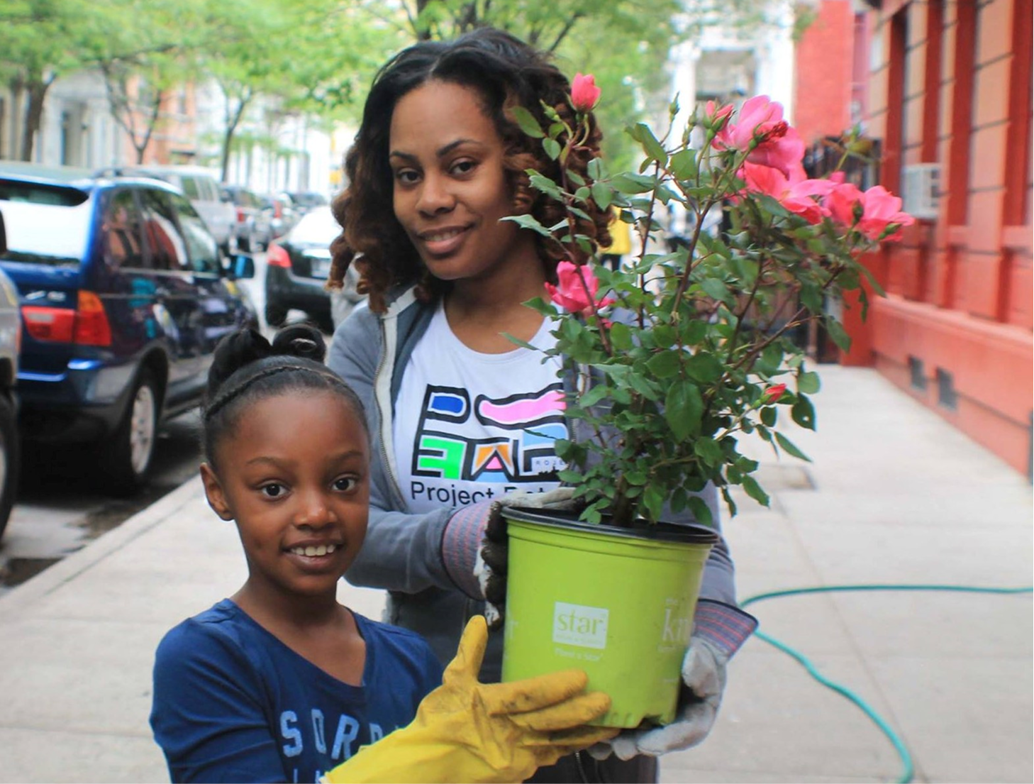 A woman and a young girl holding a potted pink flowering plant on a city sidewalk, with parked cars and buildings in the background.