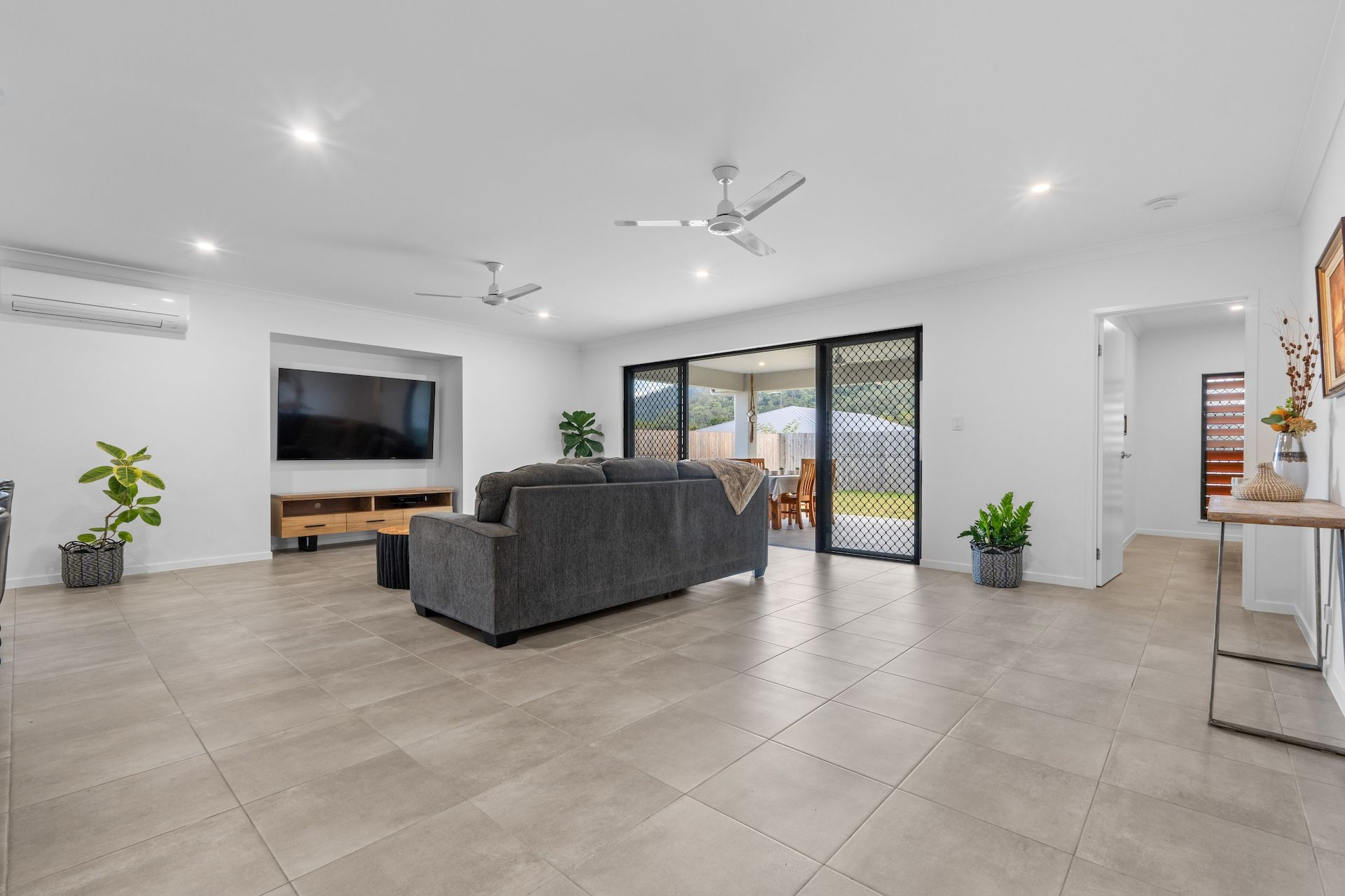 Living room with beige tiled floor, white walls, a dark gray sofa, a wooden TV stand with a flat-screen TV, potted plants, sliding glass doors leading to an outdoor patio, and decorative items on a wooden console table.