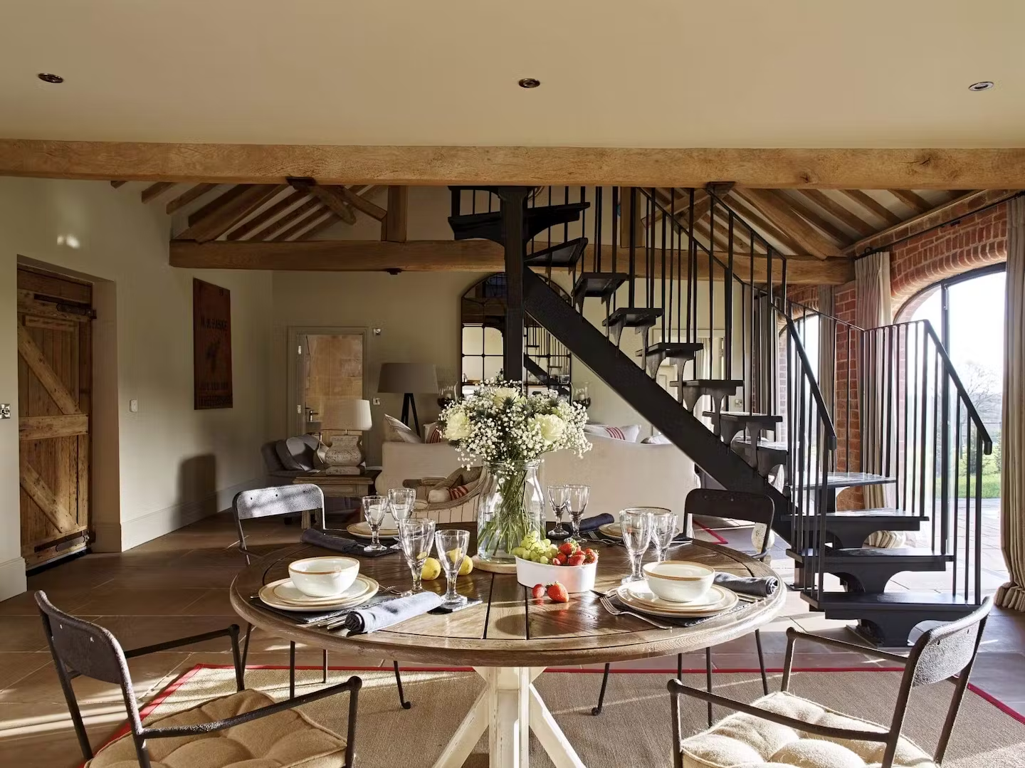 A dining room with a round wooden table set with bowls, plates, glasses, and napkins, decorated with a flower arrangement, surrounded by chairs, with a spiral staircase in the background, large windows with curtains, and a cozy seating area with a so