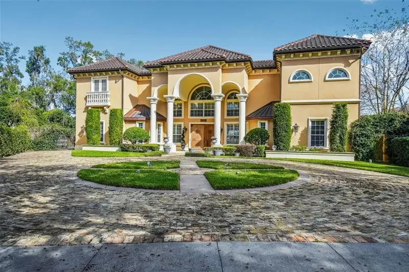Front view of a large, elegant two-story house with a curved driveway, well-maintained lawn, landscaping, and tall trees surrounding it.