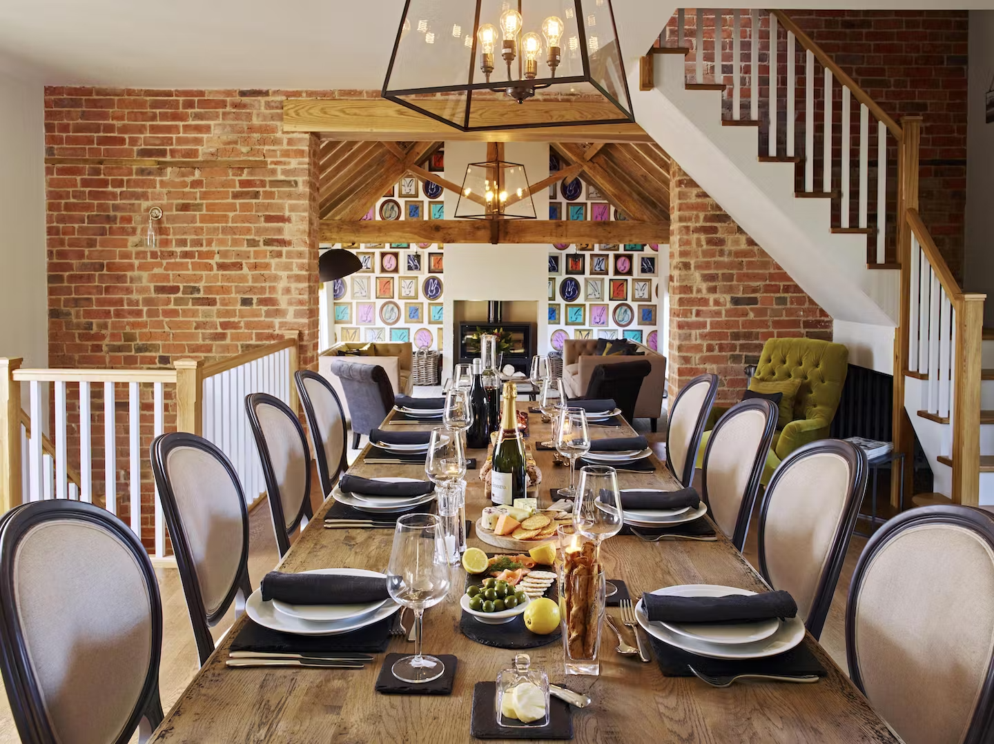 A cozy dining room with a long wooden table set for a meal, featuring plates, glasses, and black napkins. The room has exposed brick walls, wooden accents, and a staircase. In the background, there's a sitting area with colorful, framed wall decor, a