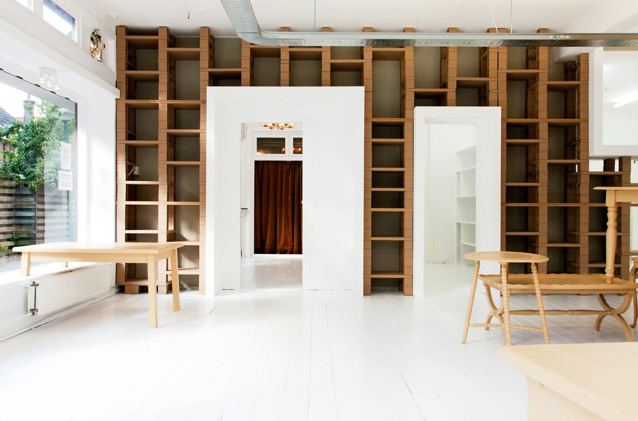 Empty room with white walls and floor, large windows on the left, and brown shelving units on the back wall. There are a few light-colored wooden tables and chairs scattered around, and a doorway with a brown curtain in the center.