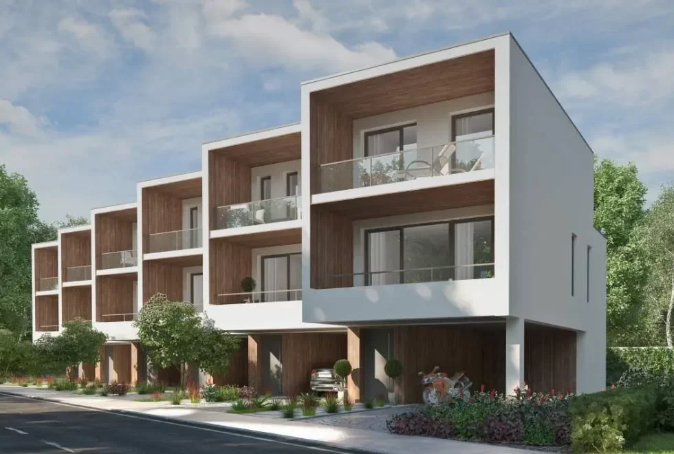 Modern multi-story residential building with white exterior walls, wooden accents, and glass balcony railings; landscaped with trees and plants.