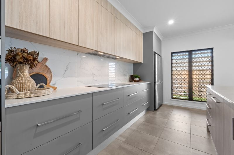 Modern kitchen with light gray lower cabinets, light wood upper cabinets, a marble backsplash, stainless steel refrigerator, and a window with black grid.
