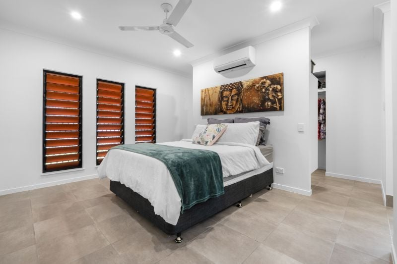 Modern bedroom with white walls, a bed with white and gray bedding, a teal blanket, and decorative pillows, three window blinds, a Buddha artwork, ceiling fan, air conditioning unit, and tiled floor.