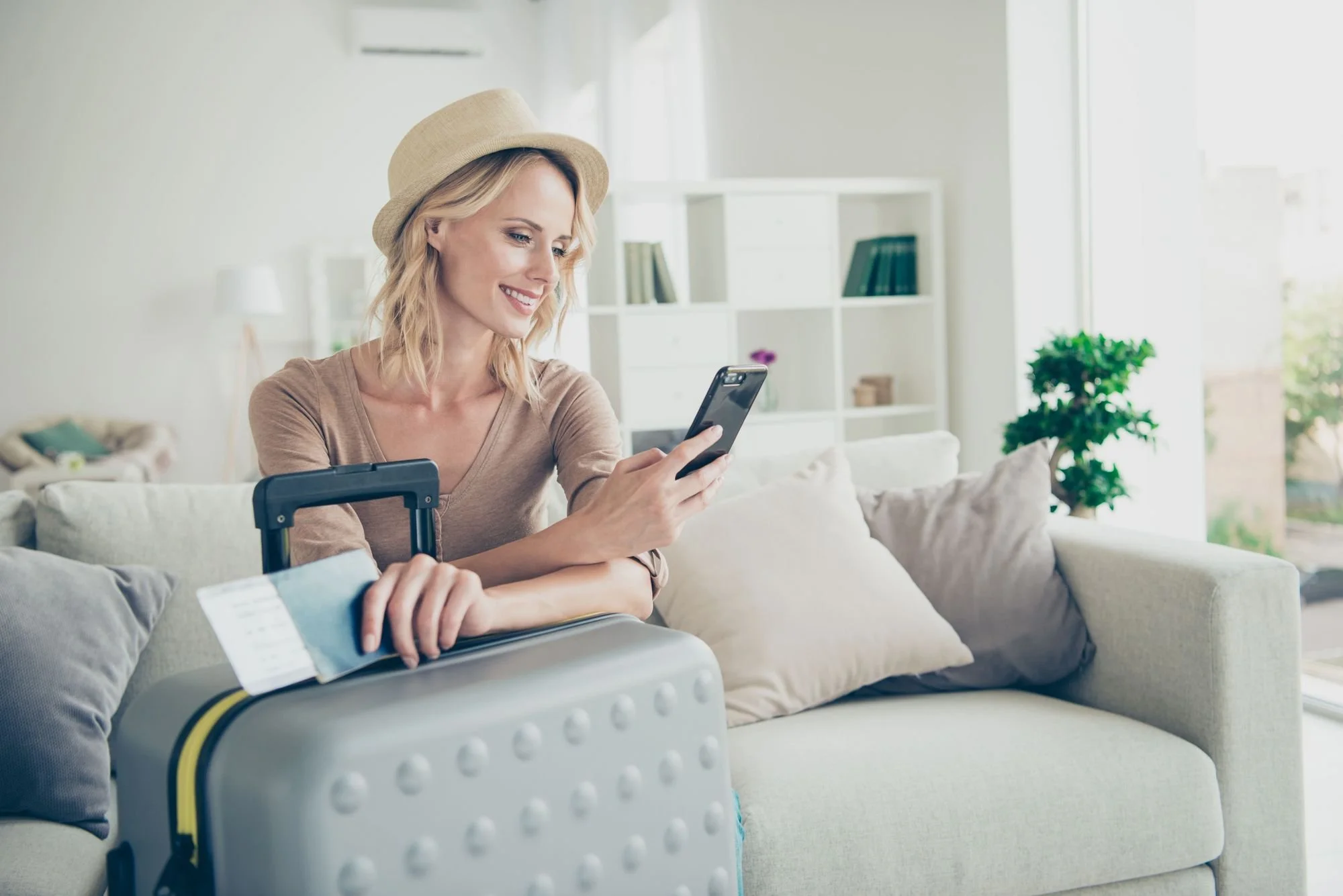 A woman sitting on a beige sofa, smiling and looking at her phone, with a silver suitcase and an airline ticket in her hands, in a bright living room with large windows and white shelves.