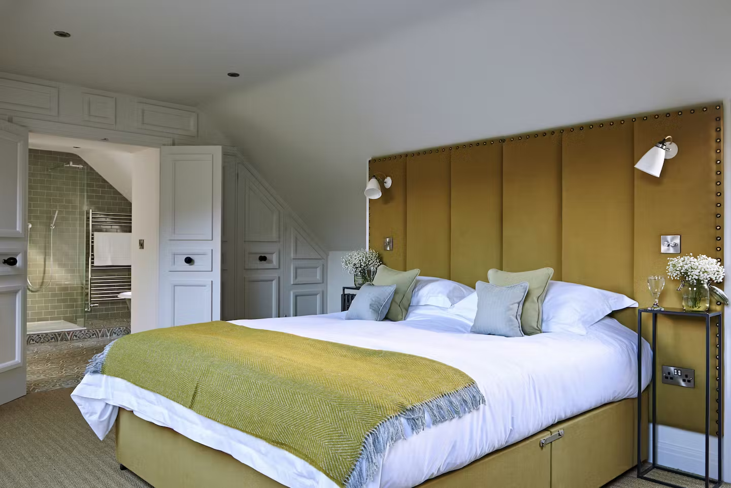 A neatly made bed with a mustard-colored headboard, white bedding, and green pillows in a modern bedroom with a visible en-suite bathroom in the background.