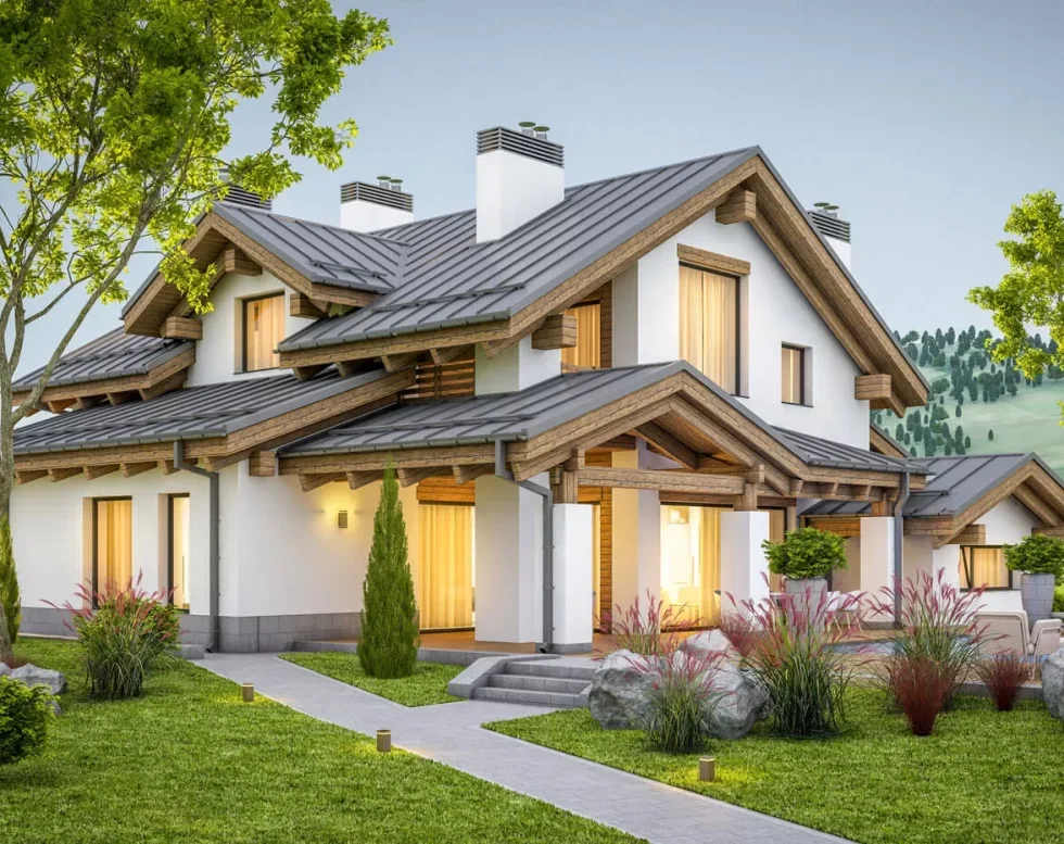 A modern two-story house with a gray metal roof and white exterior walls, surrounded by a landscaped yard with small trees, bushes, and rocks, set against a hillside with green trees.