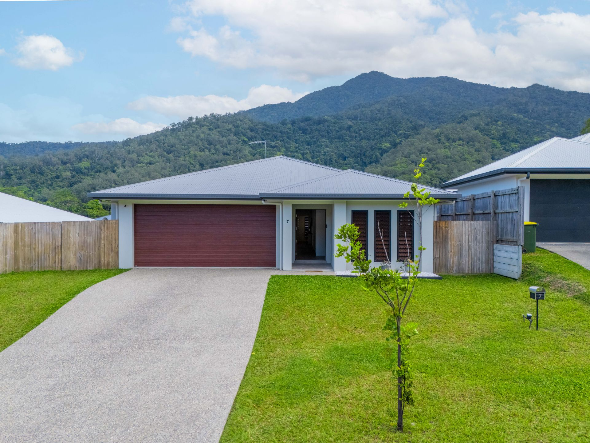 A modern single-story house with a light gray roof, dark red garage door, and white walls, situated in a grassy yard with a small tree and a mountain backdrop.