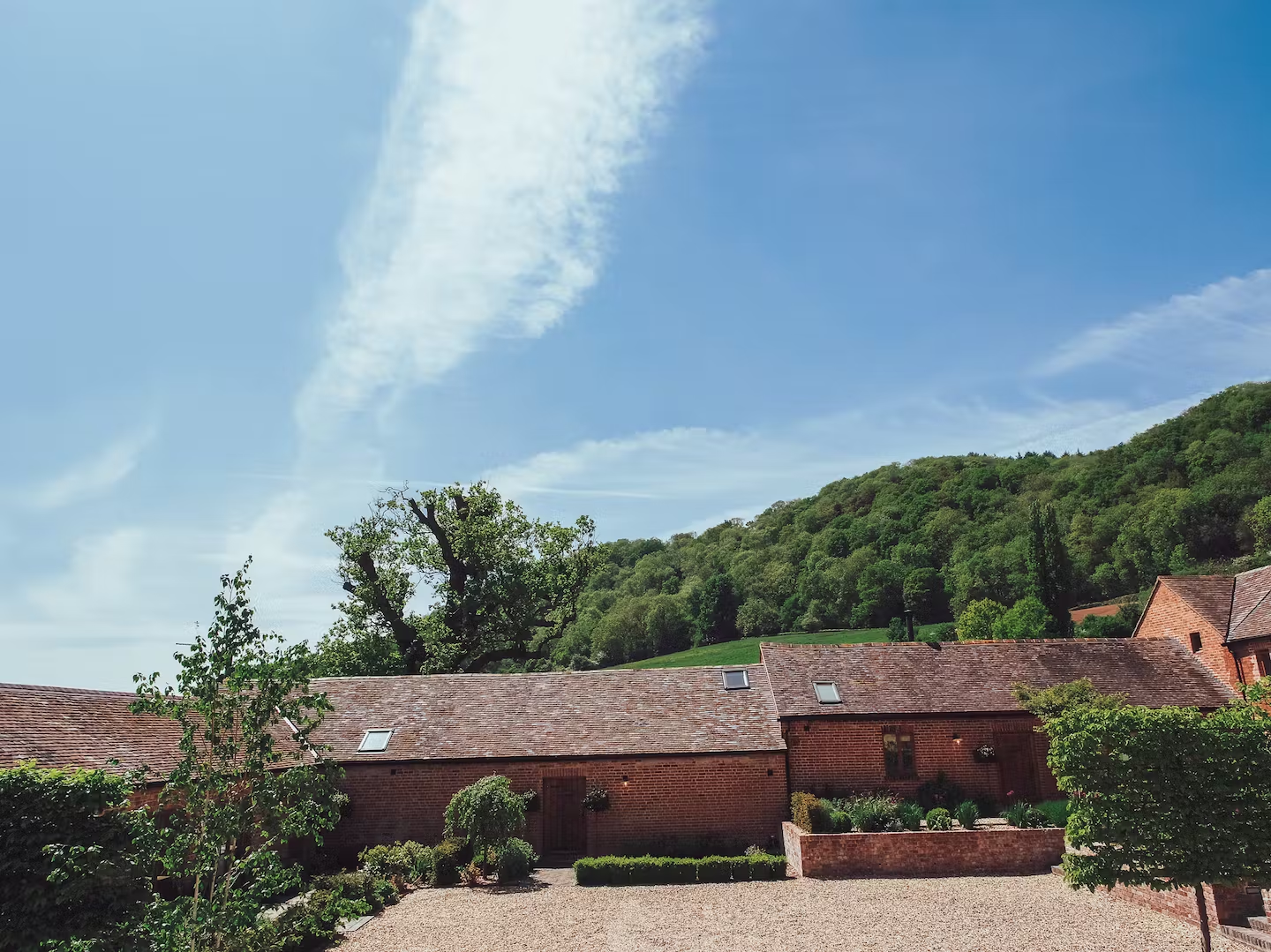 Red brick buildings with tiled roofs, surrounded by greenery, against a backdrop of a wooded hill and a partly cloudy blue sky.
