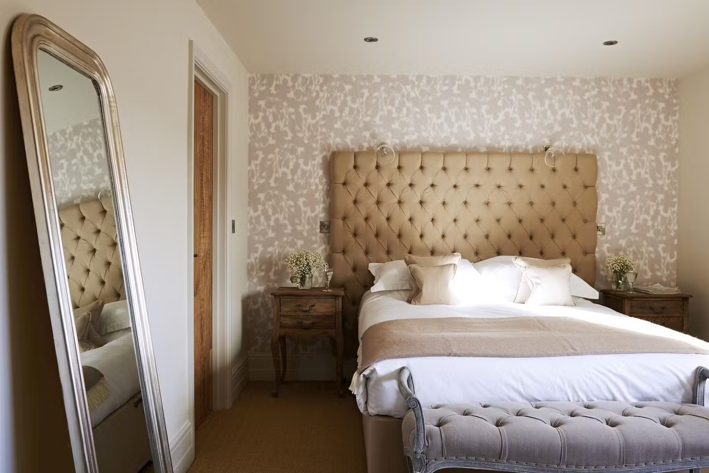 Bedroom with large tufted beige headboard, white bedding, and beige throw blanket. Two wooden nightstands with white flower vases and lamps. Decorated wallpaper on the wall, a full-length mirror leaning against the white wall, and a tufted beige benc