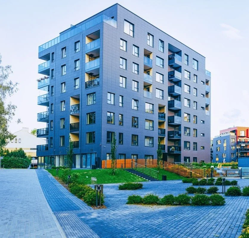 Modern multi-story apartment building with balconies, surrounded by landscaped greenery, in an urban setting.