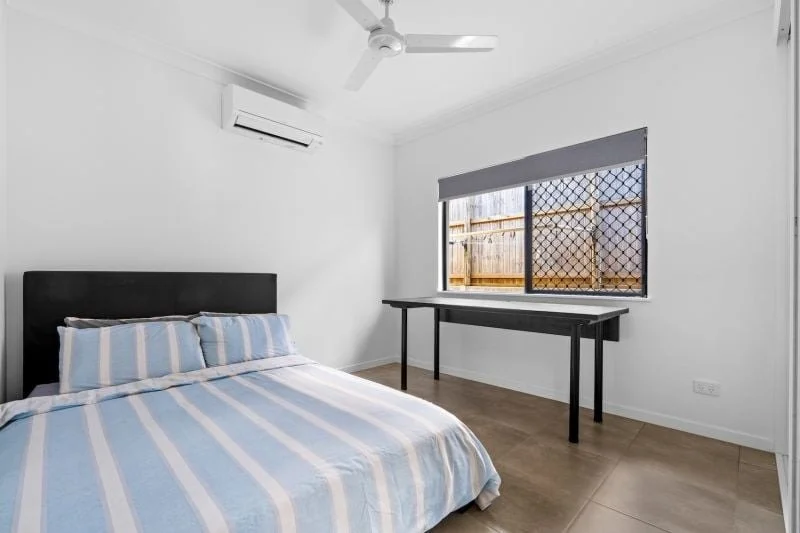 Empty bedroom with a bed, striped blue and white bedding, a window with a screen looking out to a wooden fence, a table under the window, a ceiling fan, an air conditioning unit, and wooden flooring.