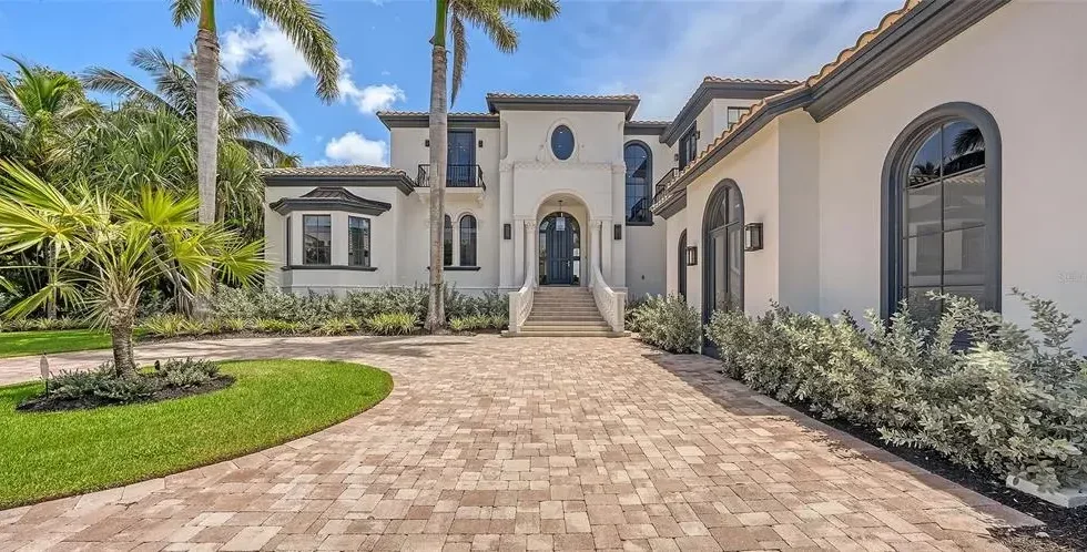 A luxurious white mansion with arched windows and a grand staircase, surrounded by palm trees and lush landscaping, with a brick driveway leading up to the entrance.