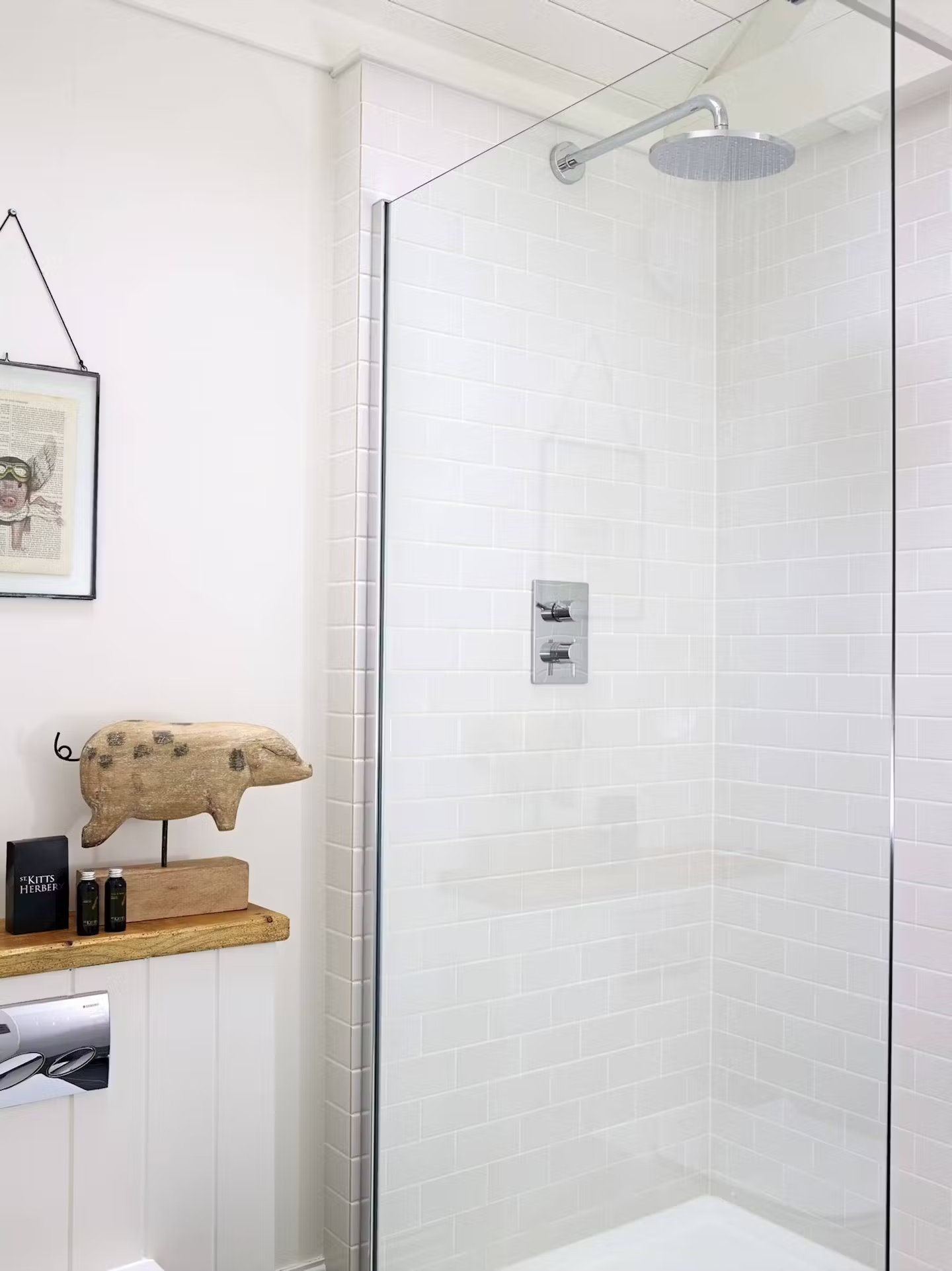 A minimalist bathroom shower with white subway tile, a black framed picture on the wall, a wooden shelf with a decorative pig sculpture, and toiletries on the shelf.