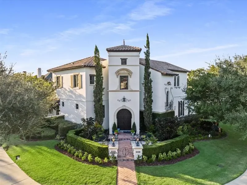 Large white mansion with a Spanish-style tile roof, arched doorway, and tall cypress trees in front, surrounded by a lush green lawn and landscaped garden.