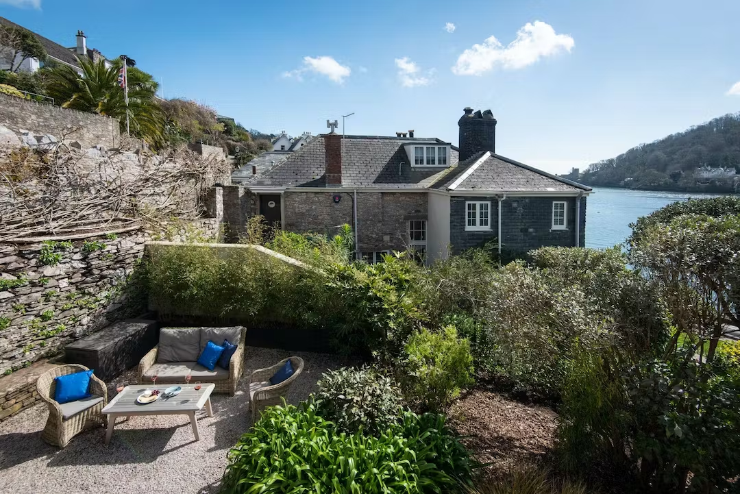 Outdoor patio with wicker chairs and a sofa with blue cushions, overlooking a house with a stone exterior and chimney, with water and hills in the background under a blue sky with scattered clouds.