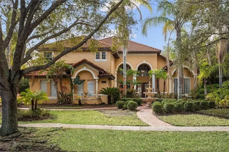 Large, two-story house with yellow stucco walls, red tile roof, and arched windows, surrounded by lush green trees and bushes, with a brick pathway leading to the front door and a decorative fountain in the yard.