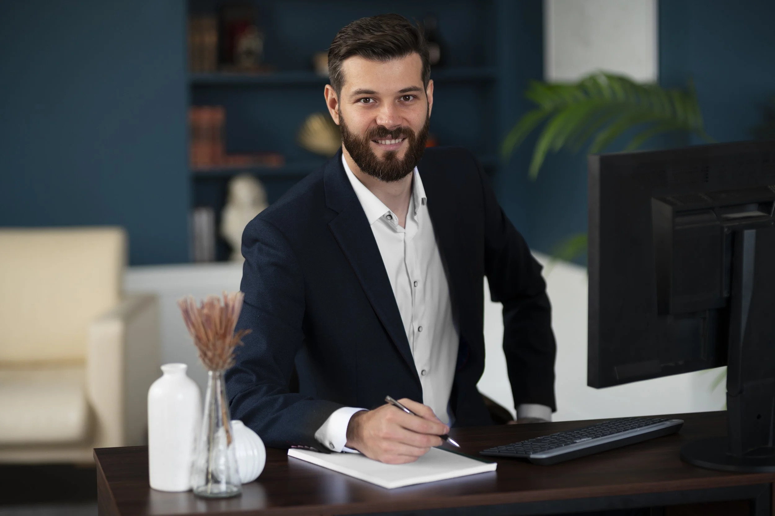 A smiling man with a beard and dark hair, wearing a navy blazer and white shirt, sitting at a desk with a notepad, pen, computer monitor, and decorative items like a white vase and a dried flower arrangement, in an office setting with blue walls and bookshelves in the background.