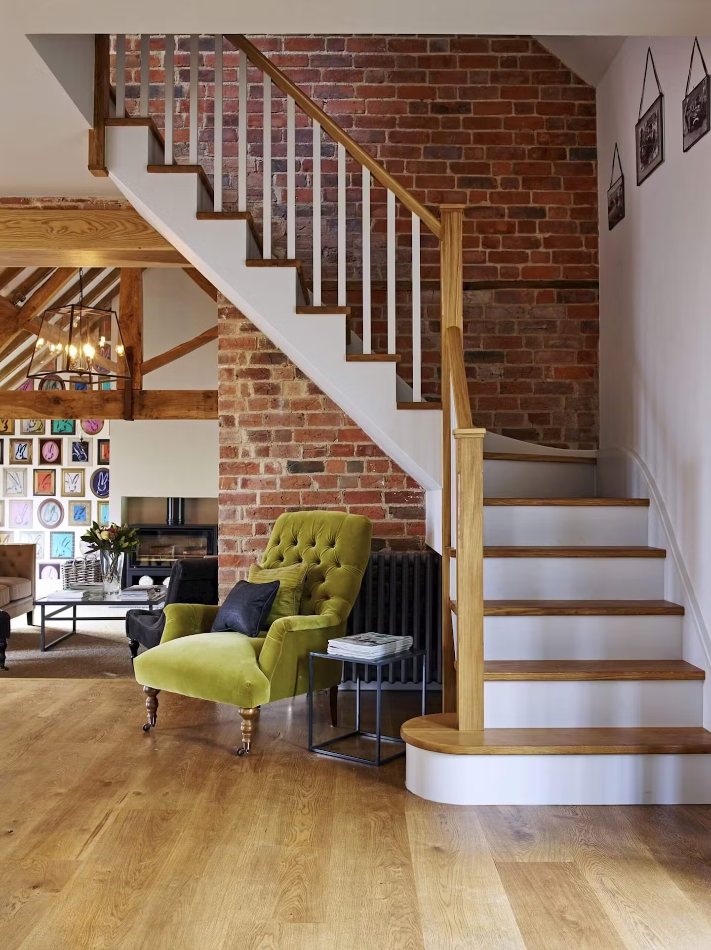 Living room with wooden floor, green upholstered armchair, black side table with magazines, brick wall behind staircase, and framed black-and-white photos on white wall to the right. Part of a dining area with patterned wall and chandelier visible in
