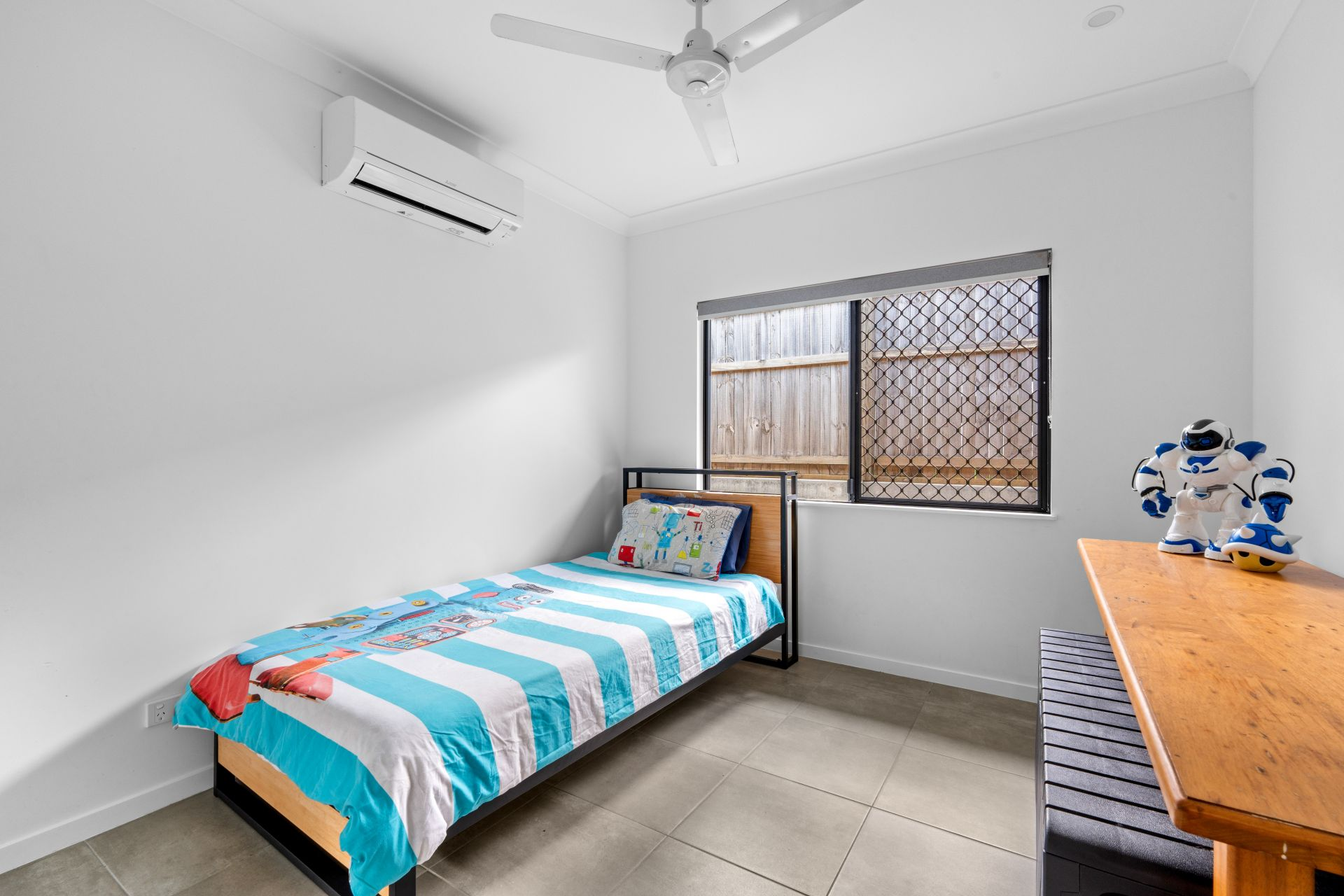 A minimalist child's bedroom with a single bed featuring a colorful bedspread and pillow, a wooden desk with robot toys, a window with a screen, and an air conditioning unit on the wall.