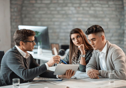 Three people in a business meeting at a conference table, discussing documents and a tablet, in a modern office setting.