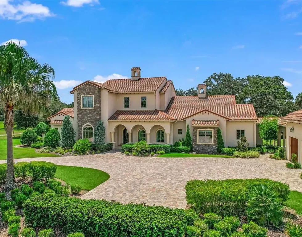 Large luxury house with a tiled roof, stone accents, and a brick driveway surrounded by lush landscaped greenery.