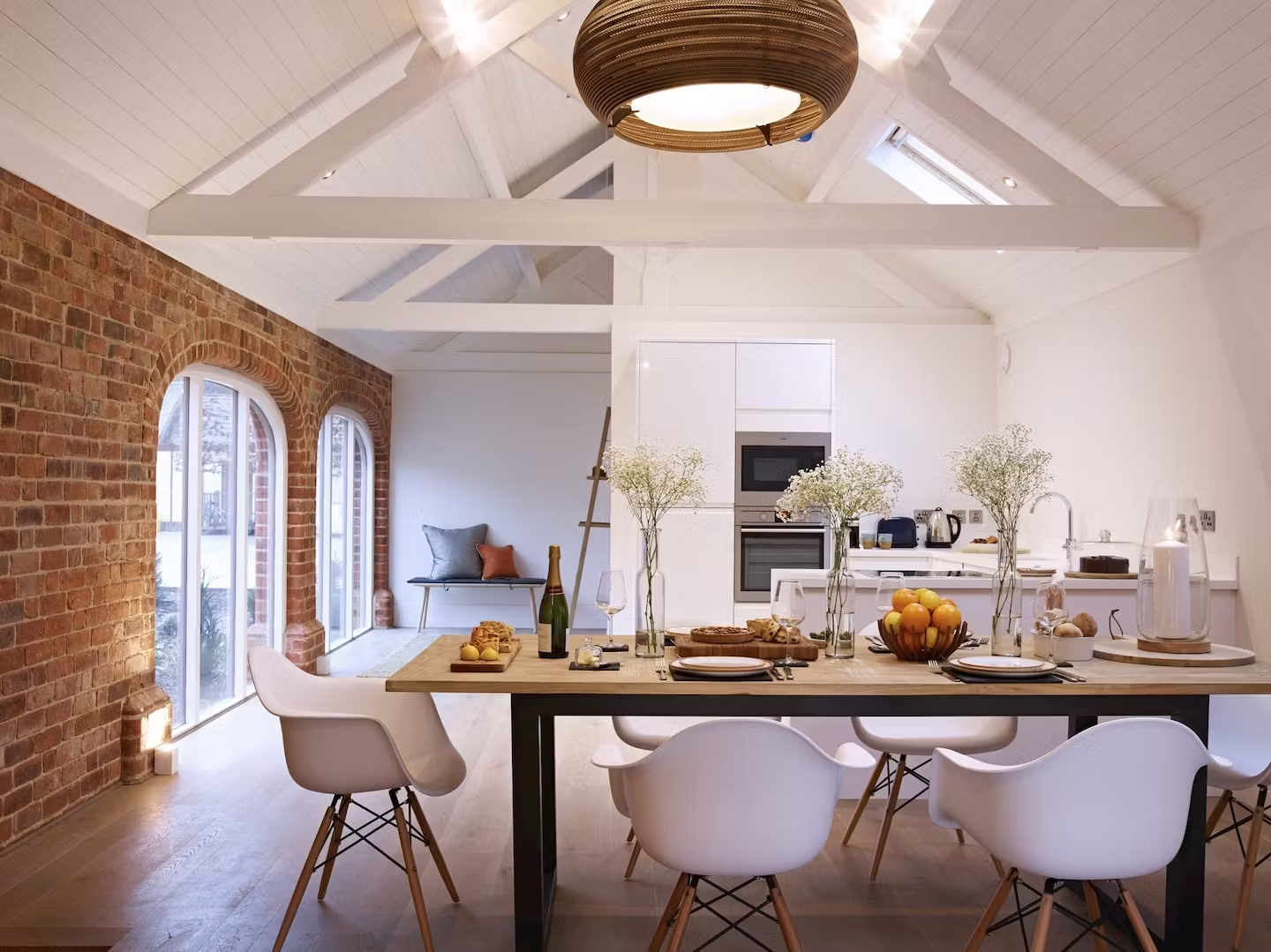 Open-concept living and dining area with exposed brick wall, large arched windows, white vaulted ceiling, and modern decor. Dining table set with plates, glasses, and centerpiece of flowers and fruit. Kitchen in background with white cabinetry and ap