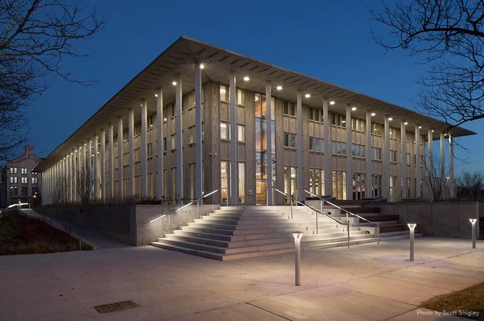 Modern building with wooden vertical panels, illuminated entrance with steps and handrails, and exterior lighting at dusk.