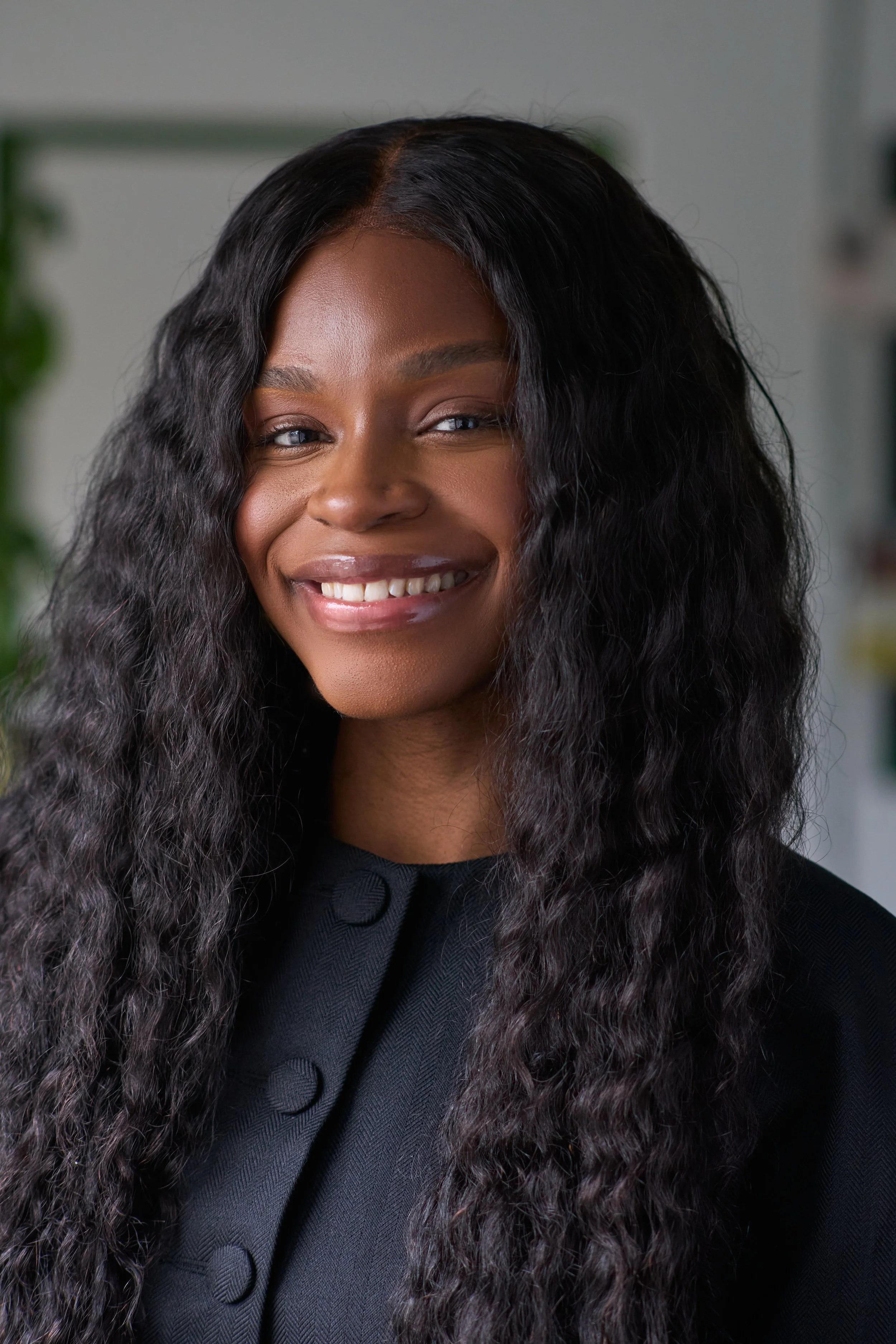 A woman with long, wavy black hair smiling, wearing a black top with a textured pattern and buttons.