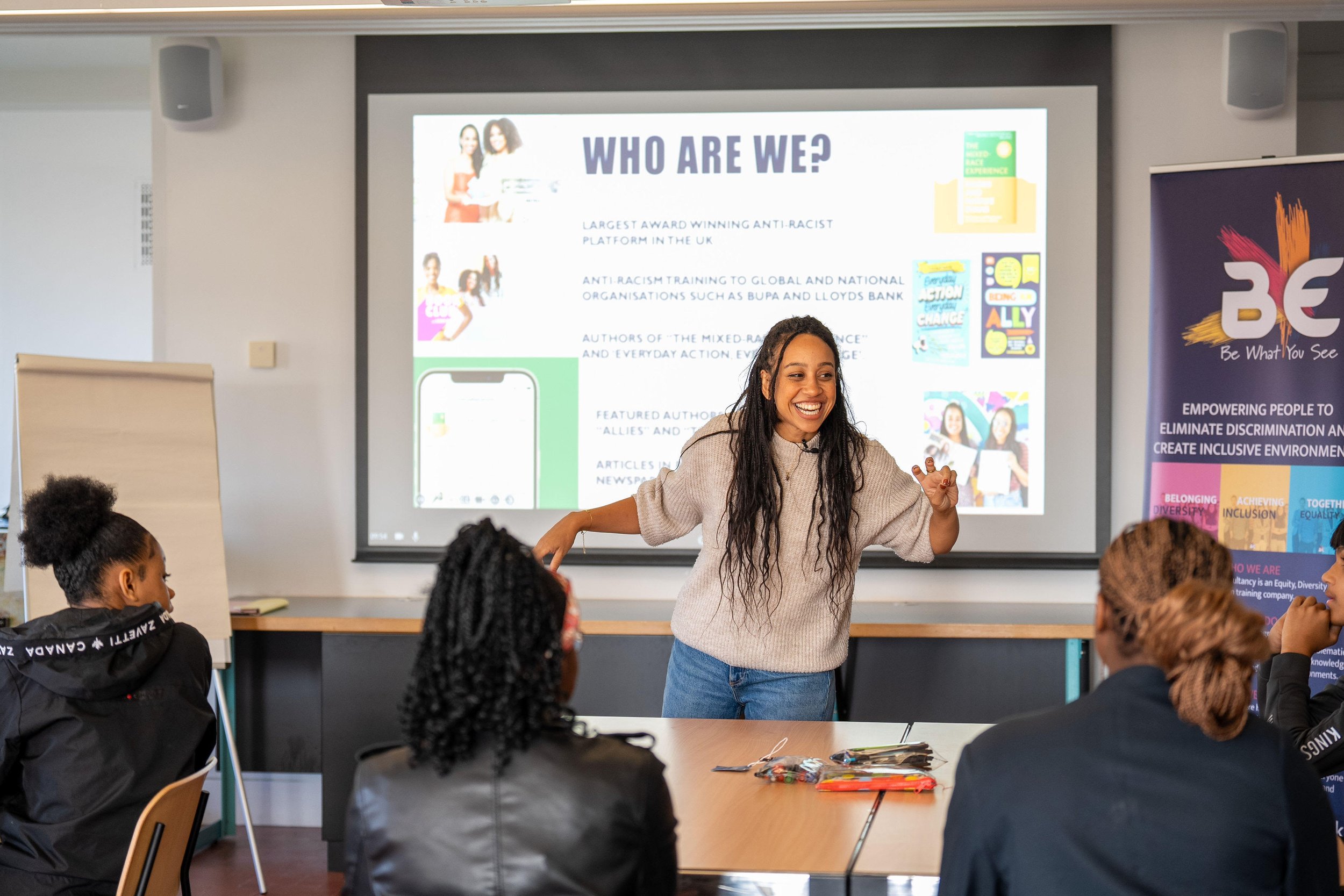 A woman giving a presentation to a group of people in a classroom. She is smiling and gesturing with her right hand, standing in front of a screen displaying a slide titled "Who Are We?" with images and text. The audience includes women listening attentively.