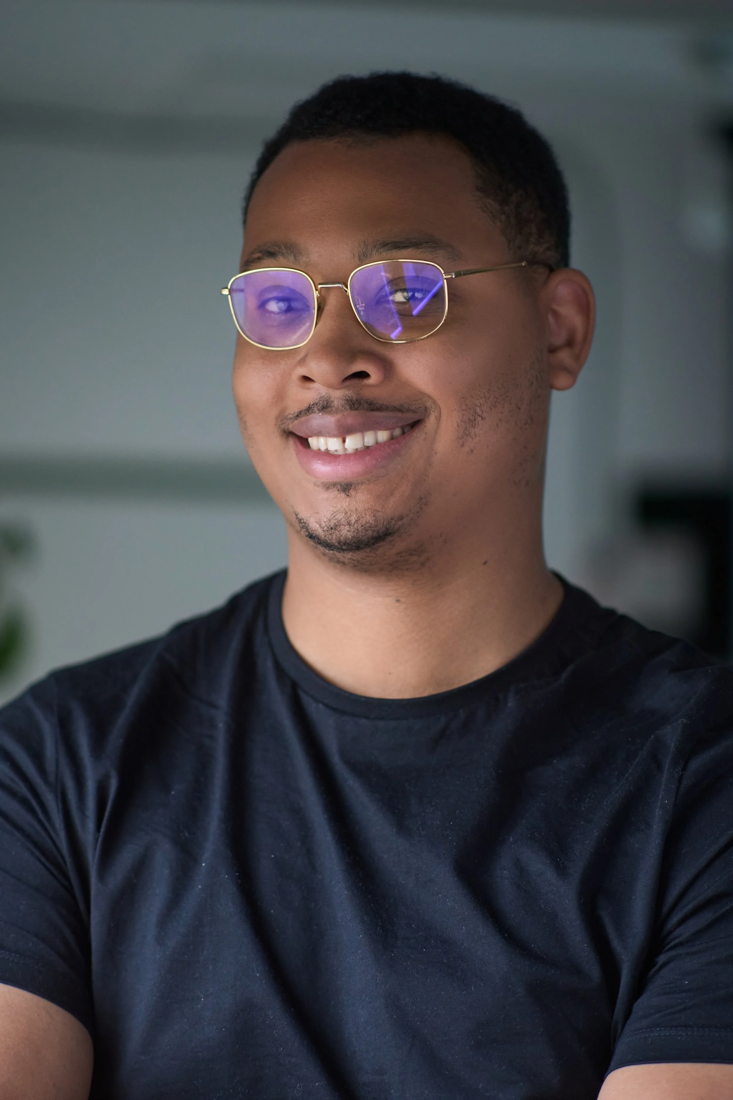 A smiling young man with short black hair, wearing glasses with purple reflection, and a black t-shirt, standing indoors.