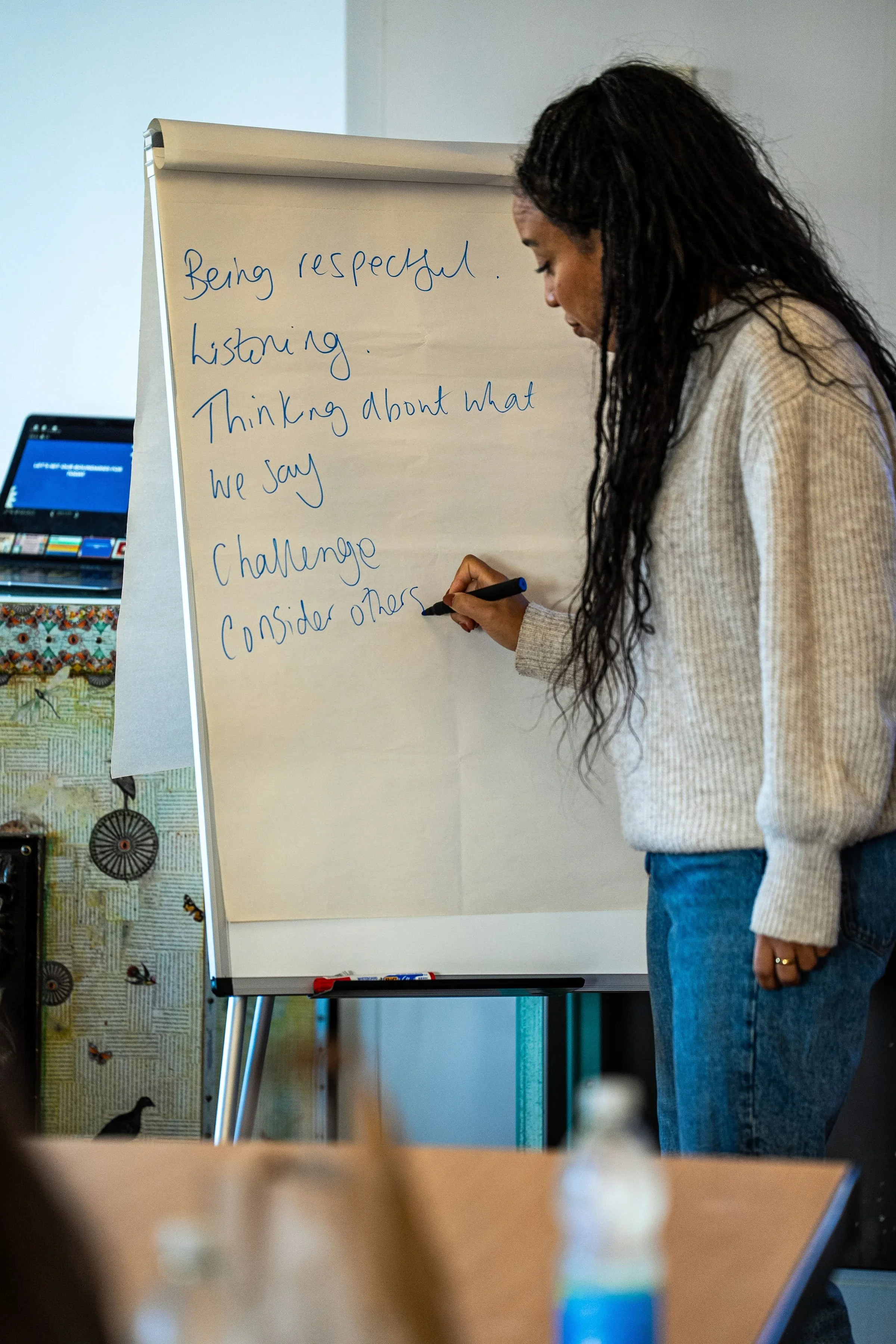 A woman with long curly hair writing on a whiteboard, with a list that includes 'Being respectful,' 'Listening,' 'Thinking about what we say,' 'Challenge,' and 'Consider others.'