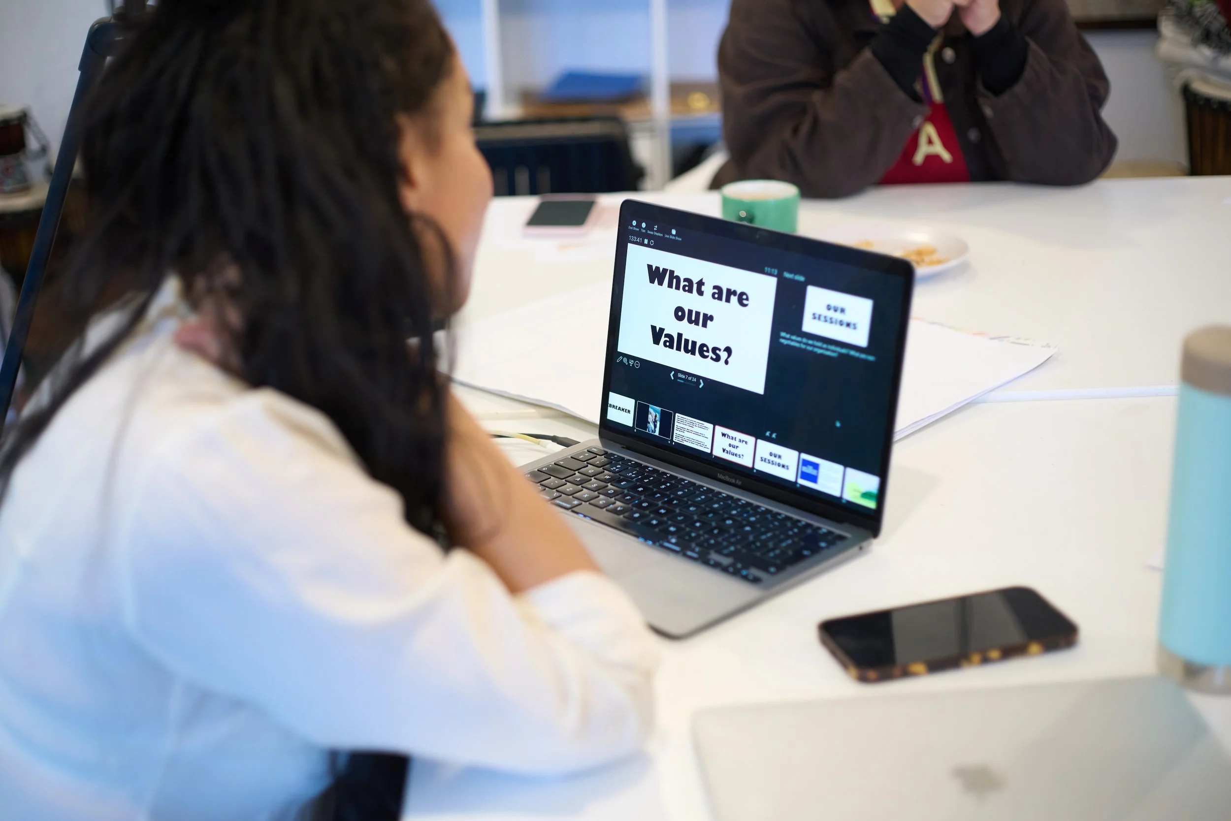 A woman sits at a white desk looking at a laptop screen that displays the question, 'What are our Values?' in bold black letters. There are papers, a smartphone, a coffee cup, and other items on the desk.