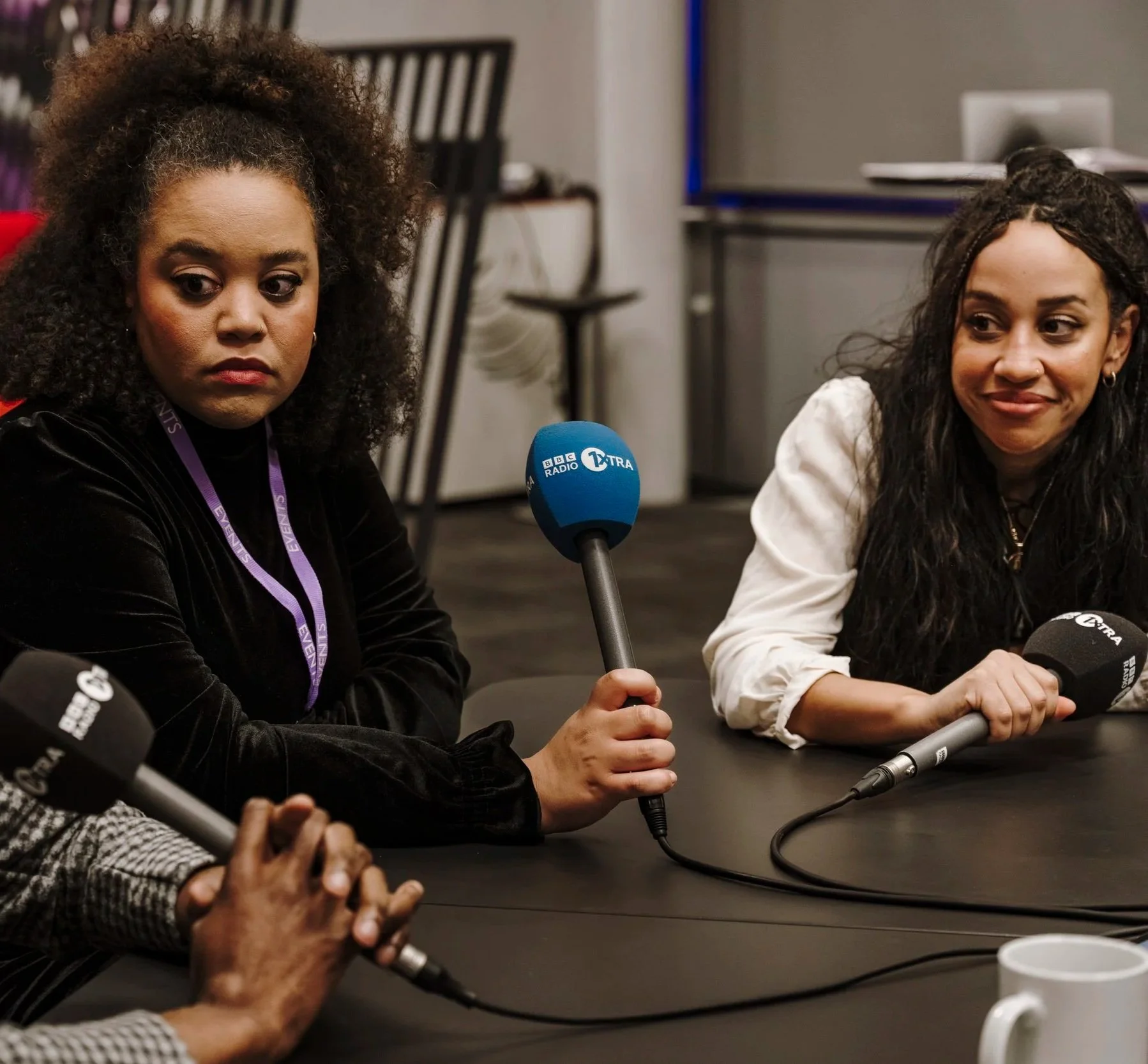 Two women sit at a table during a radio interview, holding microphones labeled BBC Radio 1 Xtra and Other Media, with a third person partially visible also holding a microphone, in a room with a black table and a bookshelf in the background.