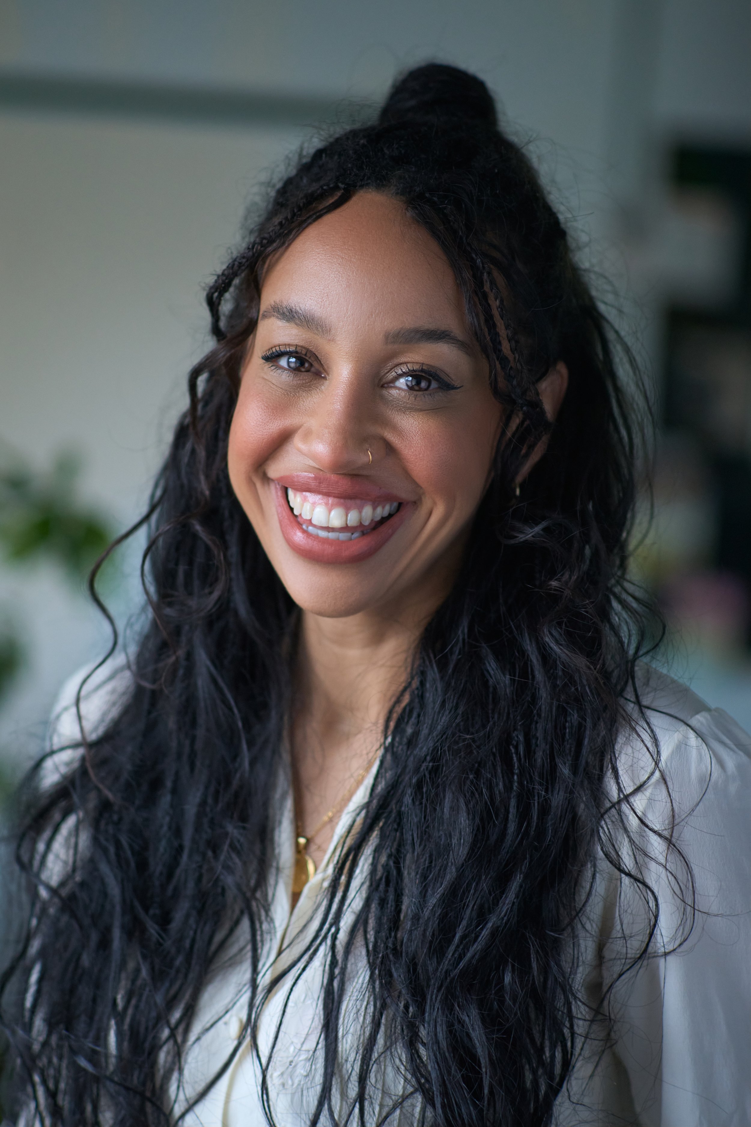 A woman with long, curly black hair styled in a half-up bun, smiling with visible teeth, wearing a light-colored blouse and a gold necklace, in an indoor setting.