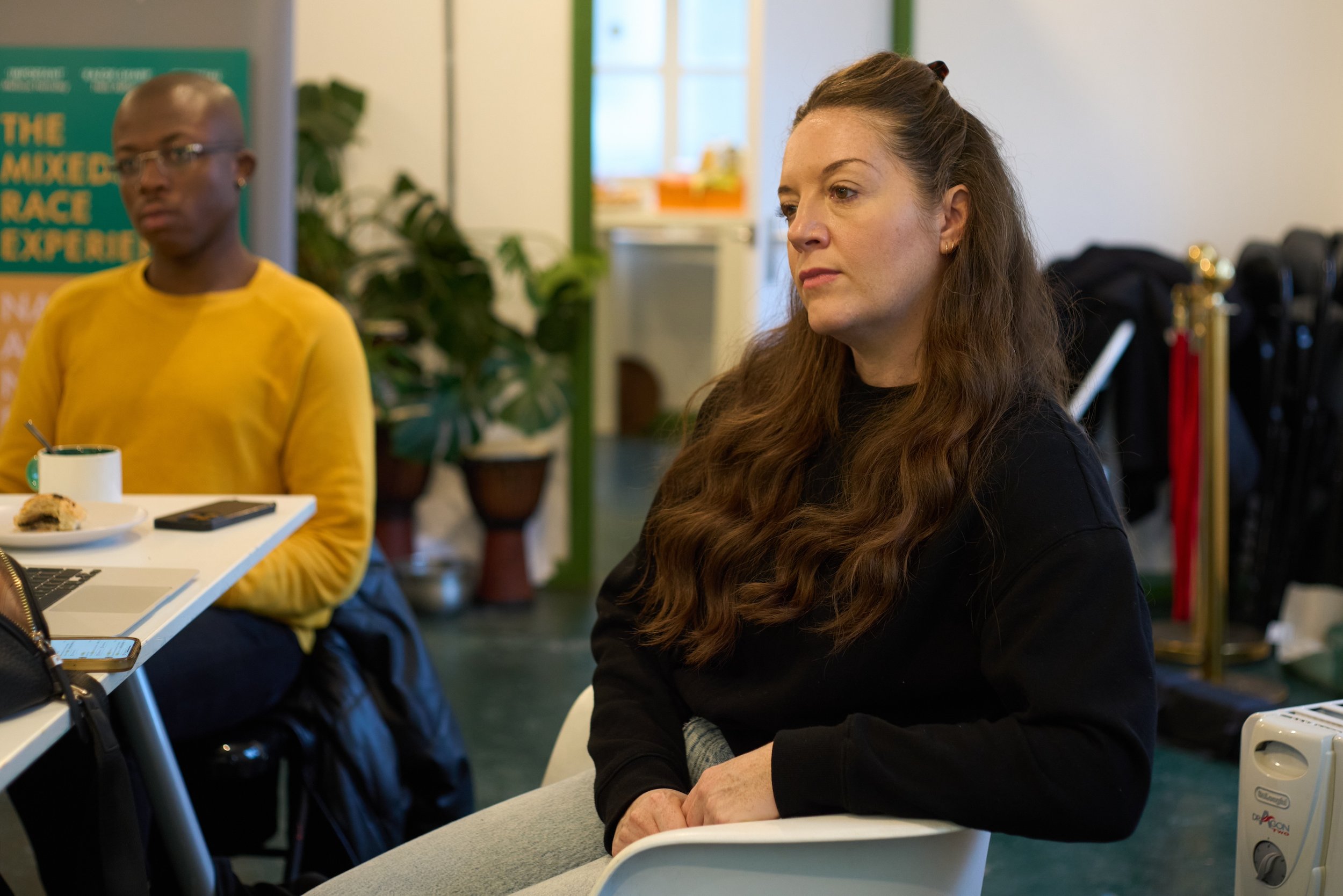 A woman with long, wavy brown hair, wearing a black shirt, sitting attentively in a white chair in a meeting or classroom setting. In the background, a man with glasses and a yellow shirt is sitting at a table with a plate of food, a cup, and a phone. Some plants and a green poster are also visible.
