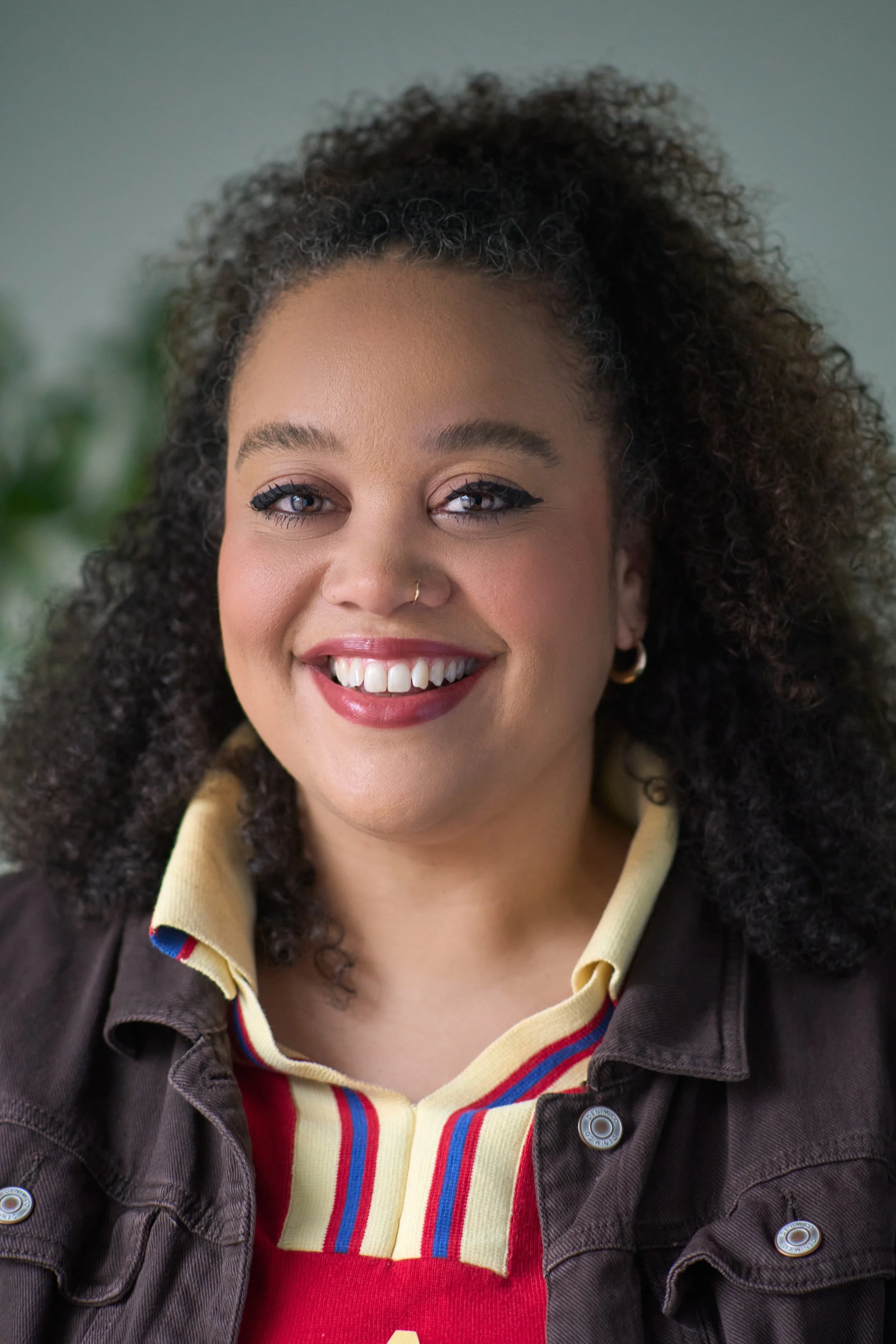 A smiling woman with curly hair, gold hoop earrings, and a nose ring, dressed in a colorful striped shirt and dark jacket.