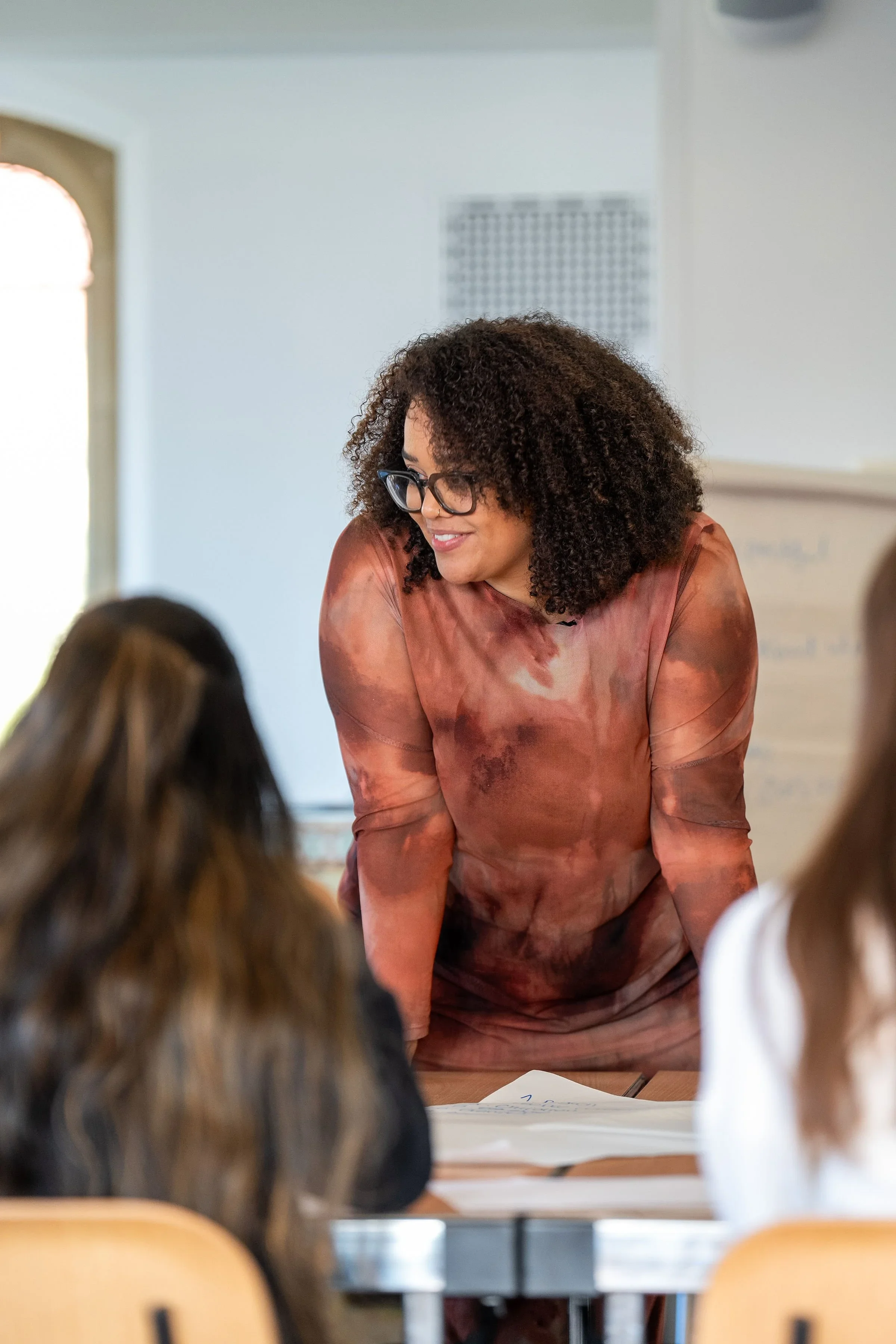 A woman with curly hair and glasses is leaning over a table, smiling during a class or workshop, with students' heads visible in the foreground.